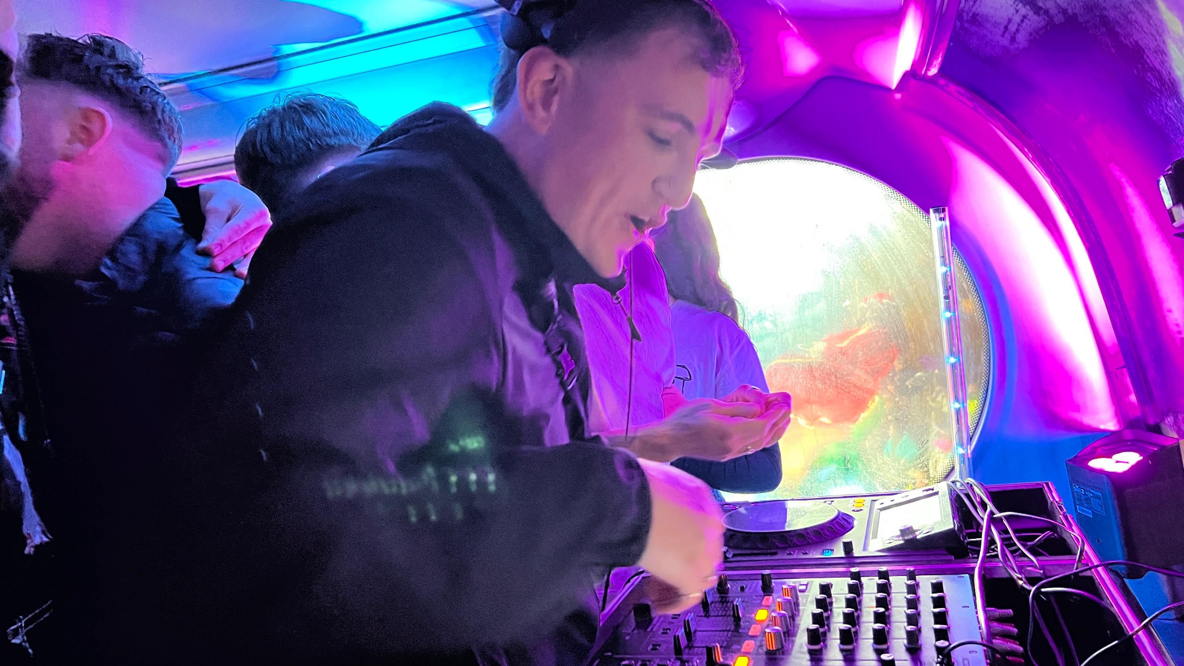 DJ at the decks at the front of the bus, lit in blue and purple lights.