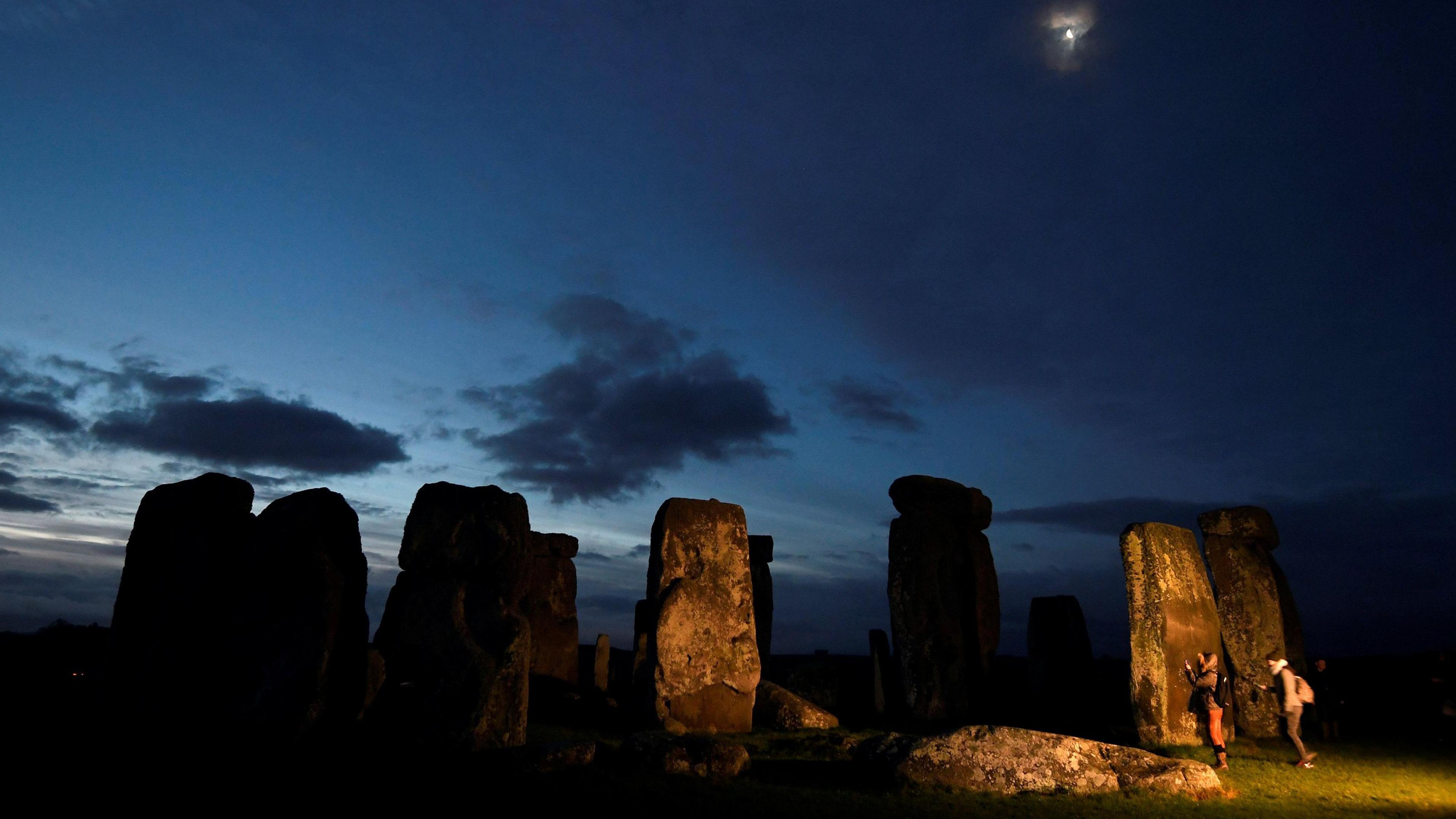 Stonehenge at dawn, with a mainly dark sky with glimmers of brightness and the moon overhead and two people next to the standing stones.