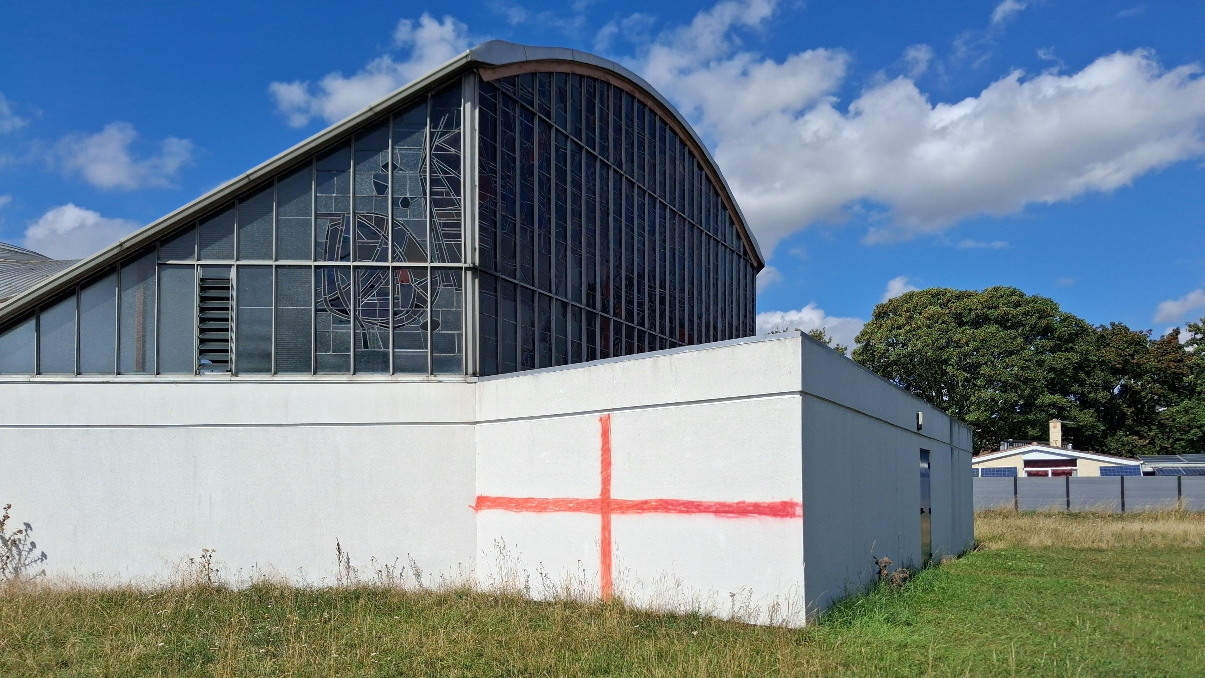 There is a white building with windows above. There is a red cross painted on a white wall. There is also long grass and trees in the distance.