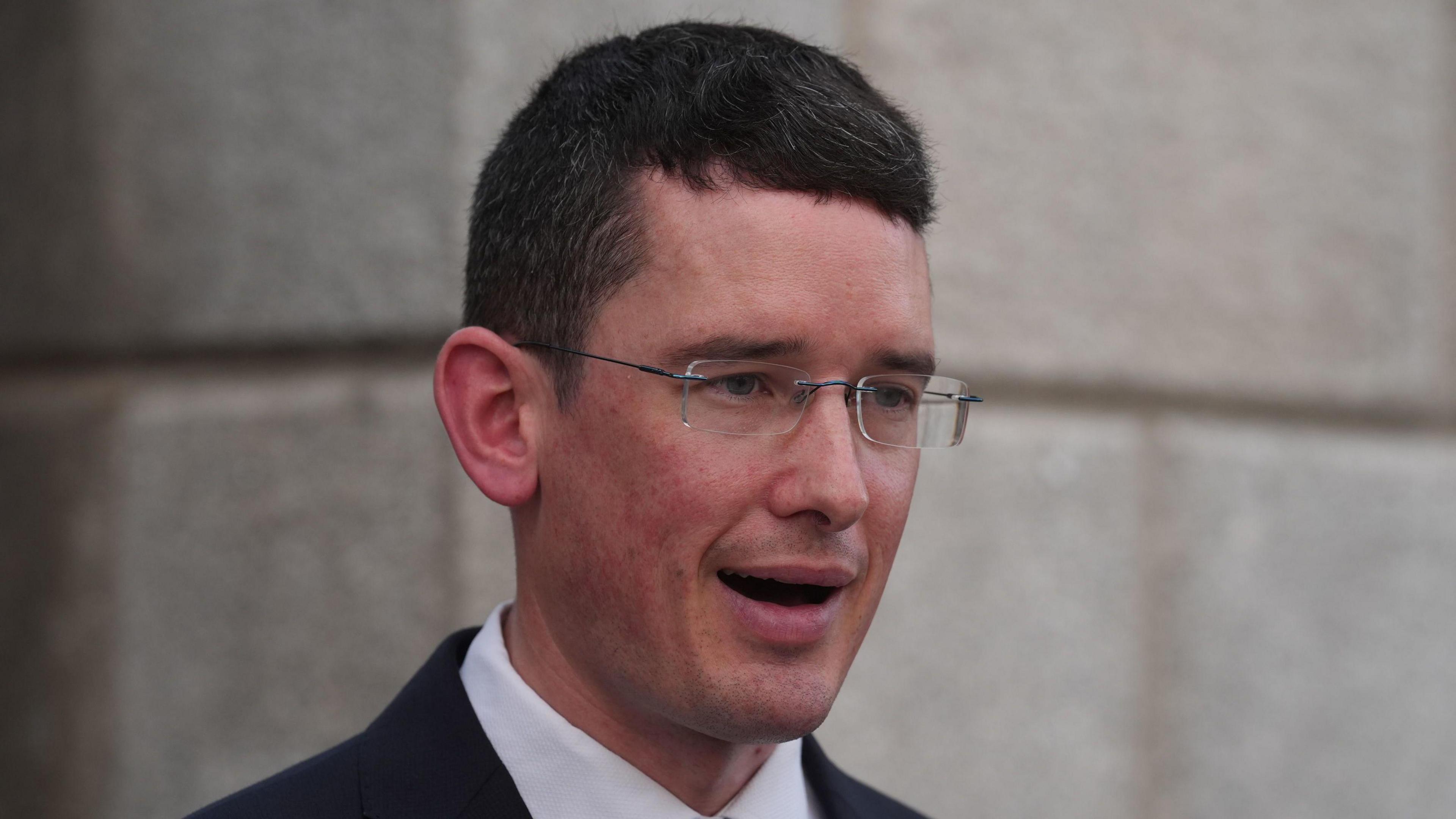 Enoch Burke looking to the right, his mouth is open as if he is speaking. He has dark hair and glasses.