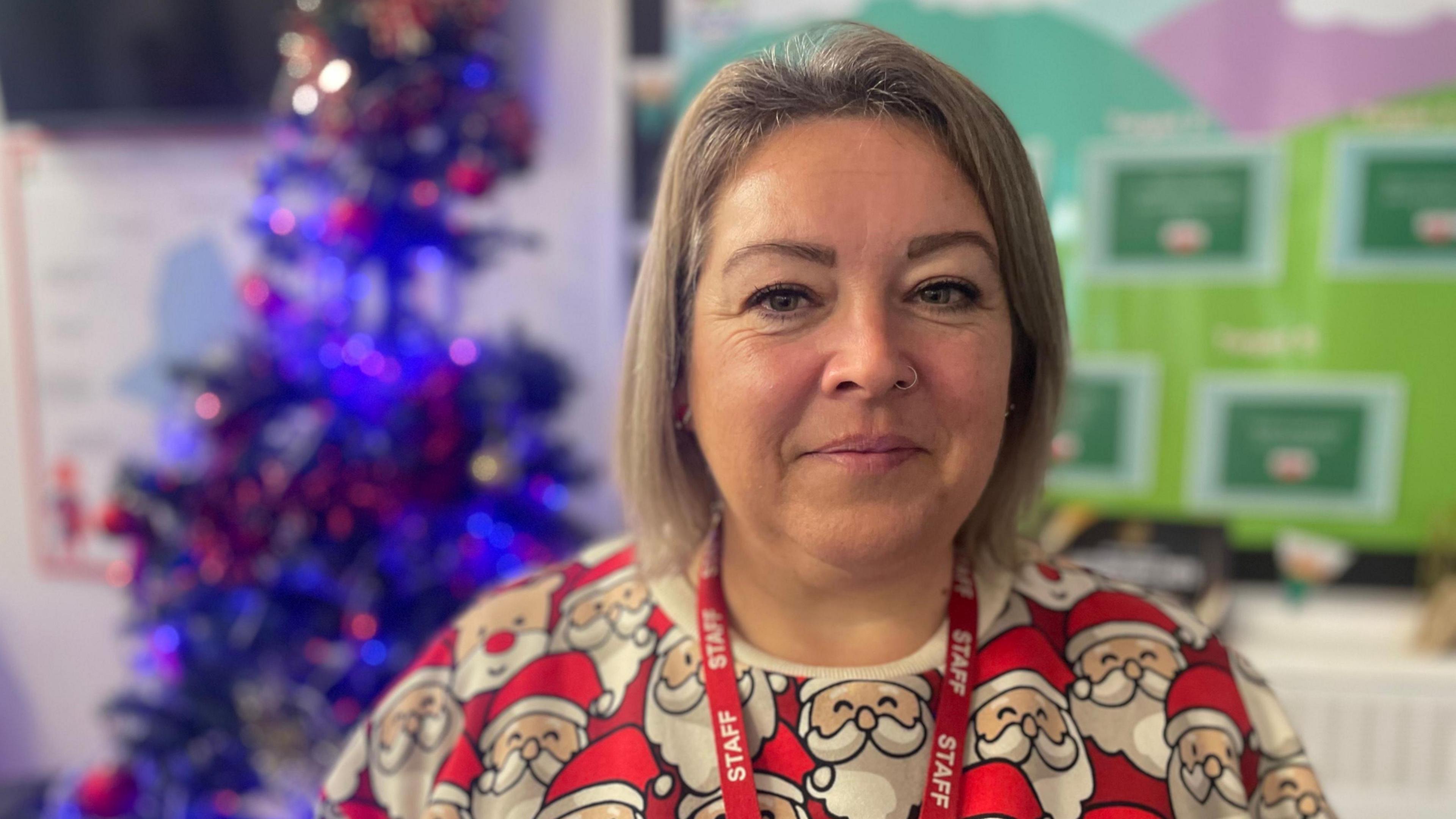 Angharad Evans, sy'n gynorthwy-ydd dysgu gyda gwallt byr golau yn gwisgo siwmper Nadolig gyda choeden Nadolig yn y cefndir