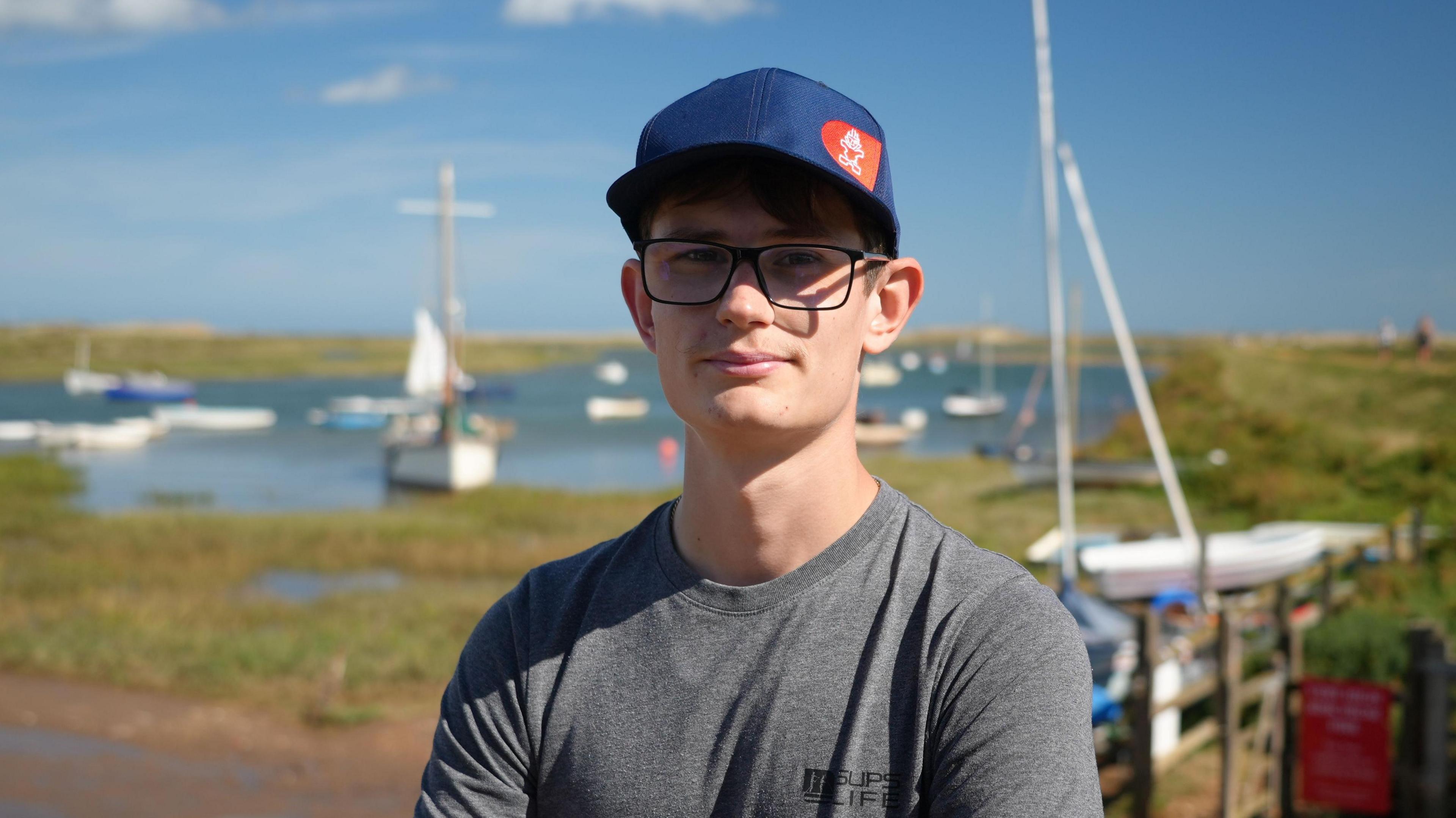 Charlie Wagg back on dry land at Burnham Overy Staithe in Norfolk. He is wearing a blue cap, glasses and grey t-shirt with water and marshes behind.
