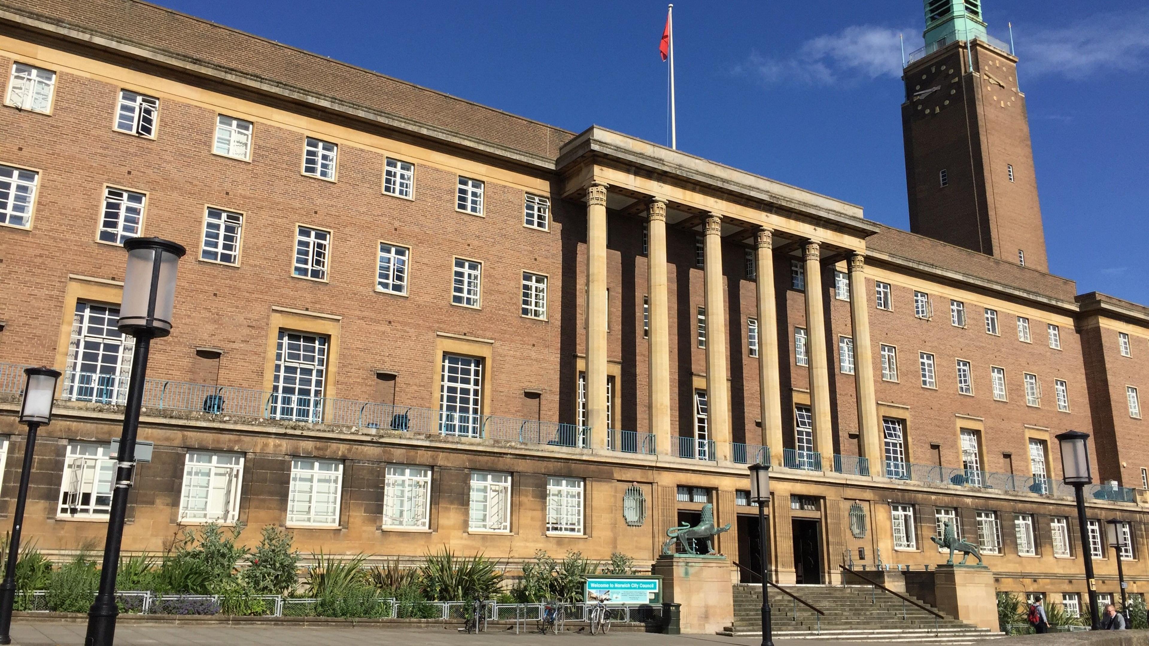 A city hall is shown with a clock tower climbing high above the building to the right of the image, with dozens of windows shown across the building's four floors and steps to the front leading to the pavement.