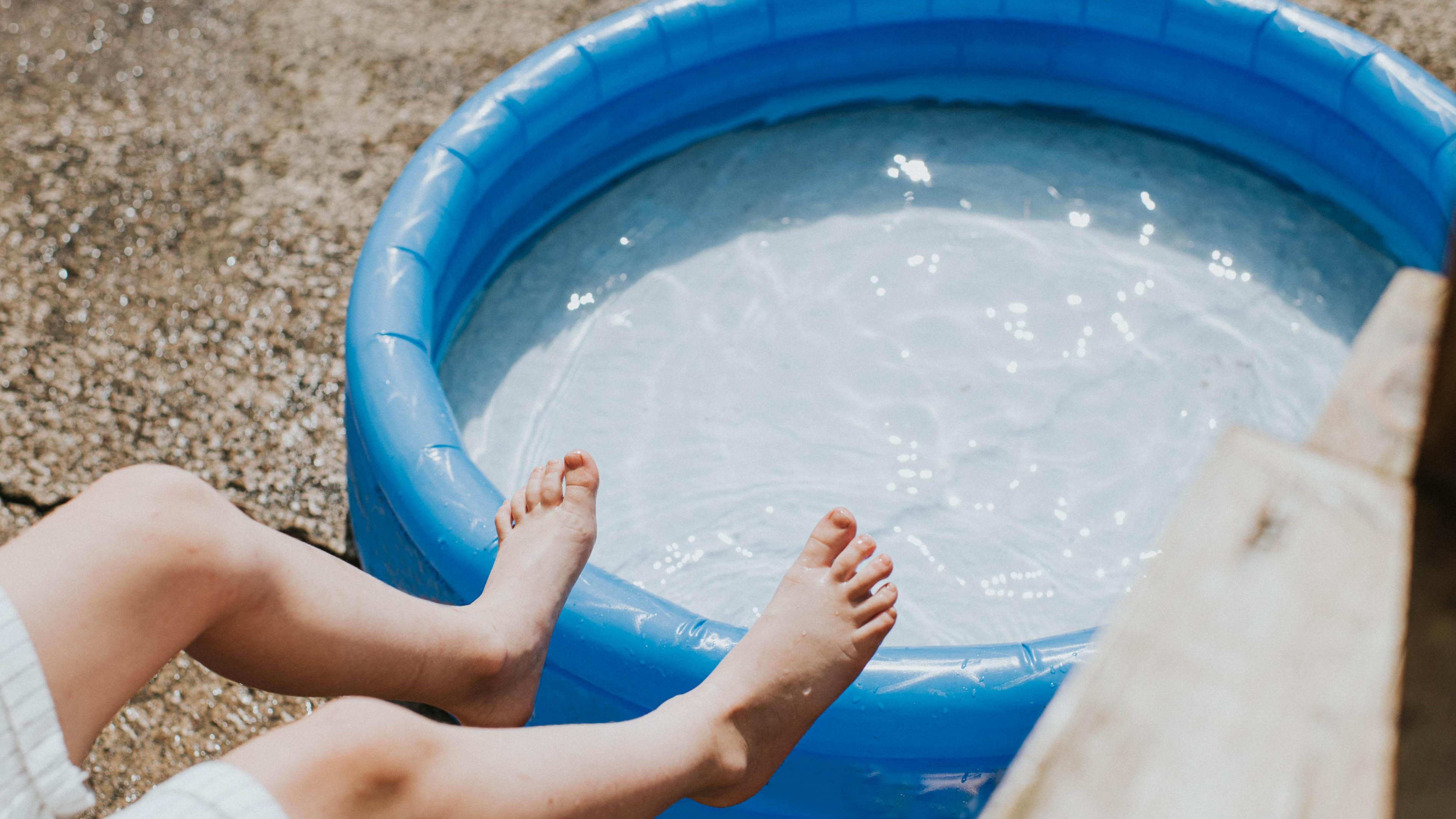 A pair of feet rest against a full paddling pool during hot sunshine.