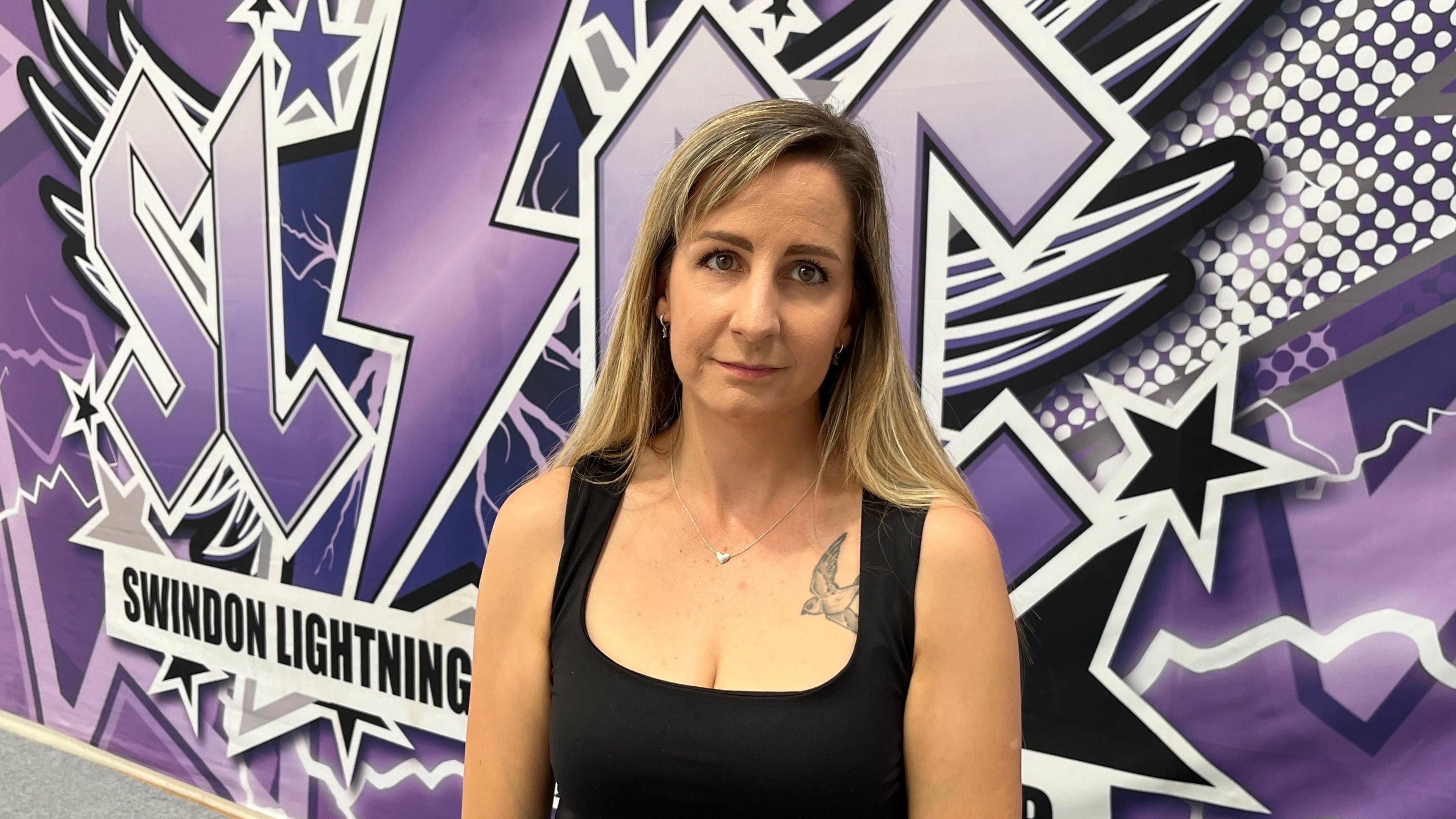 Swindon Lightning Cheerleading club director Millie Fannin pictured with long blonde hair and a black vest top standing in front of a purple, black and white back drop with the Swindon Lightning Logo on it.