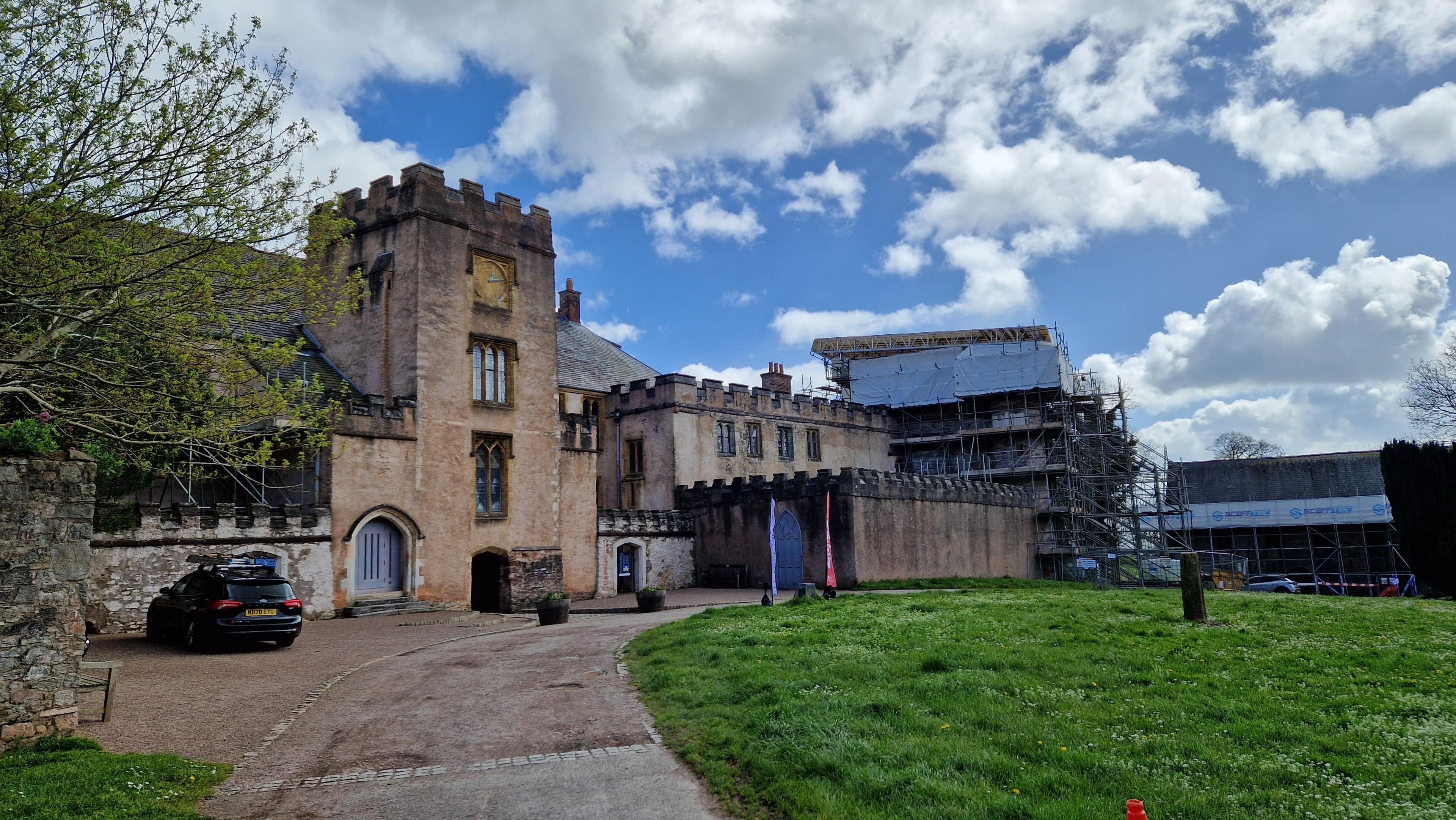 The photo shows branches of a tree on the left-hand side which has green leaves. Next to it is Torre Abbey, which has turrets of a castle in the middle and along the roof of the building. There is a green space on the right-hand side of the photo which is part of the golf course.