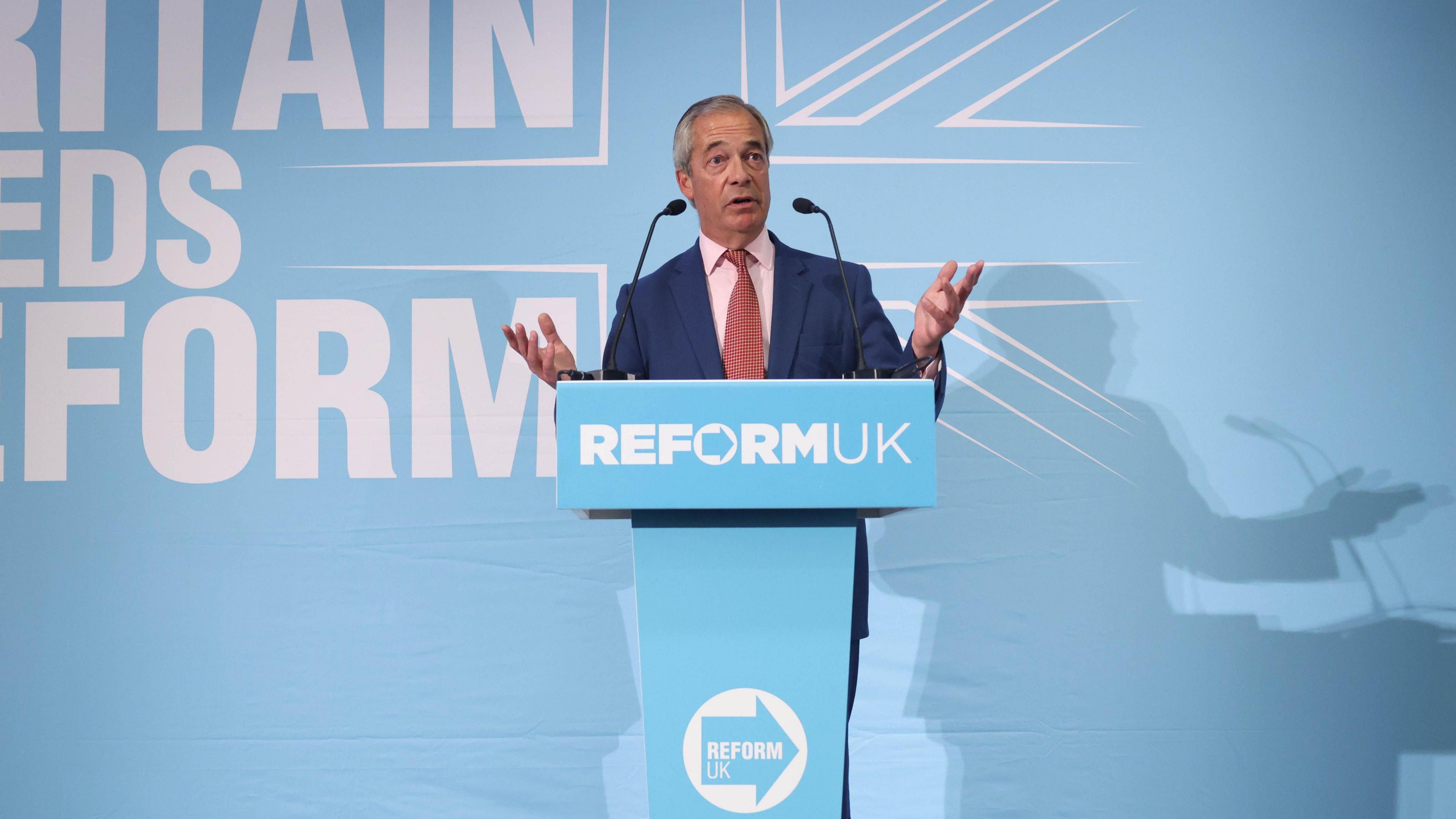 Nigel Farage, a man with grey hair in a blue suit and a pink shirt and a red tie. He's standing behind a pale blue podium that says Reform UK on it. He's in front of a background which says Britain Needs Reform.