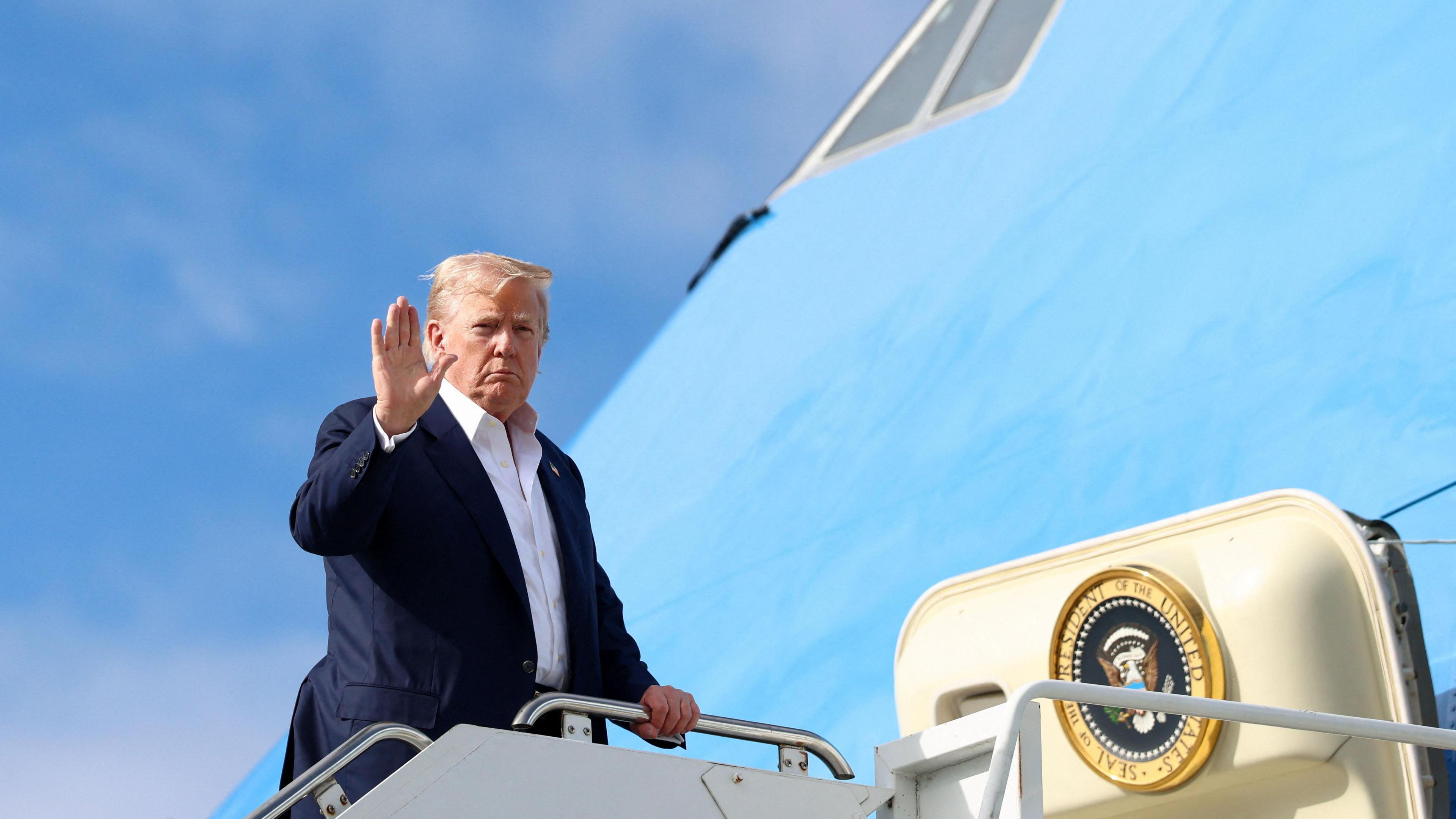 President Trump, in dark blue suit and open-necked white shirt, waves as he boards his plane. The sky is blue.