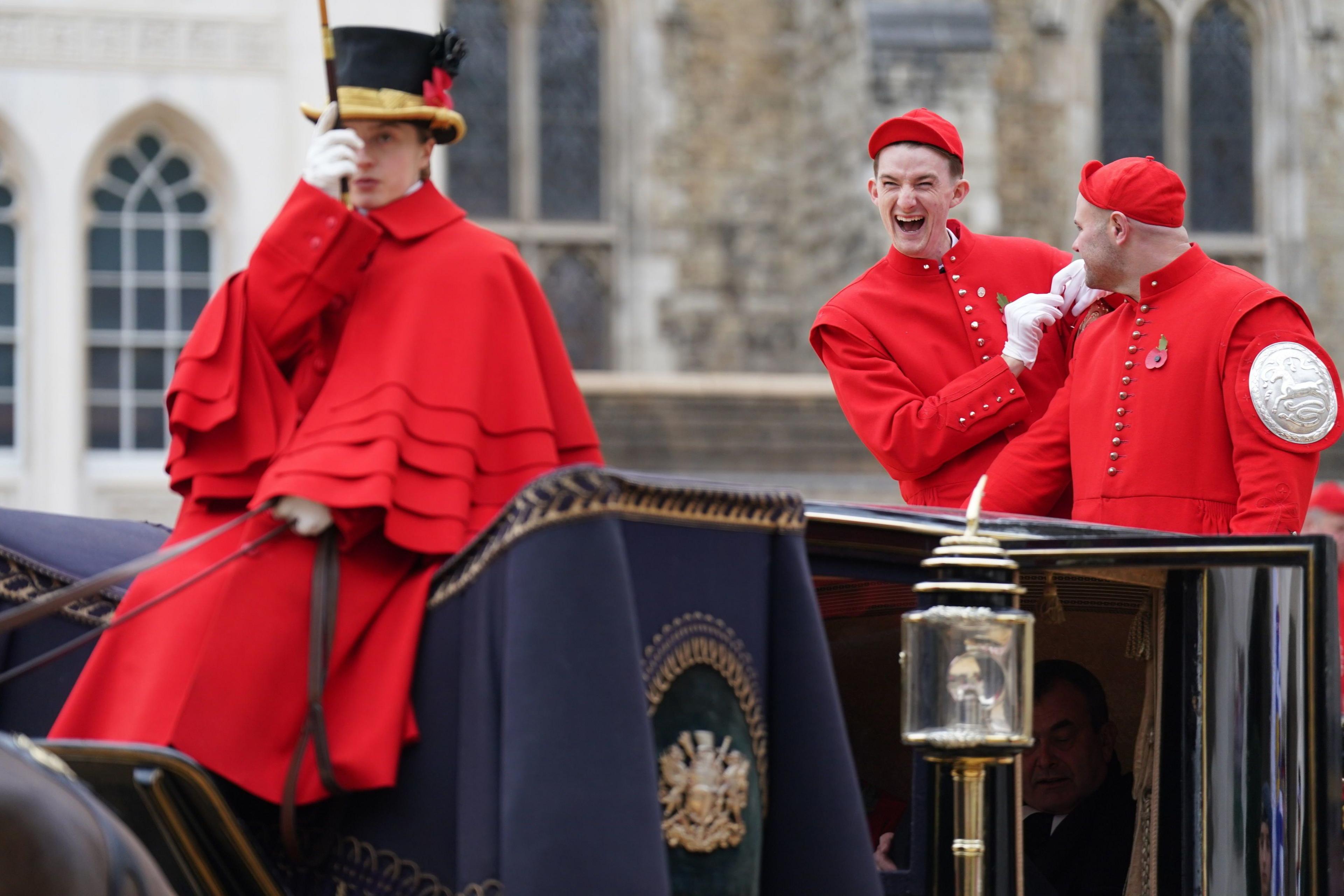 Two men in red uniforms with caps share a light-hearted moment during the Lord Mayor's Show, while standing next to a ceremonial carriage. A third person in red, wearing a black top hat with a feather, sits in the coach box. 