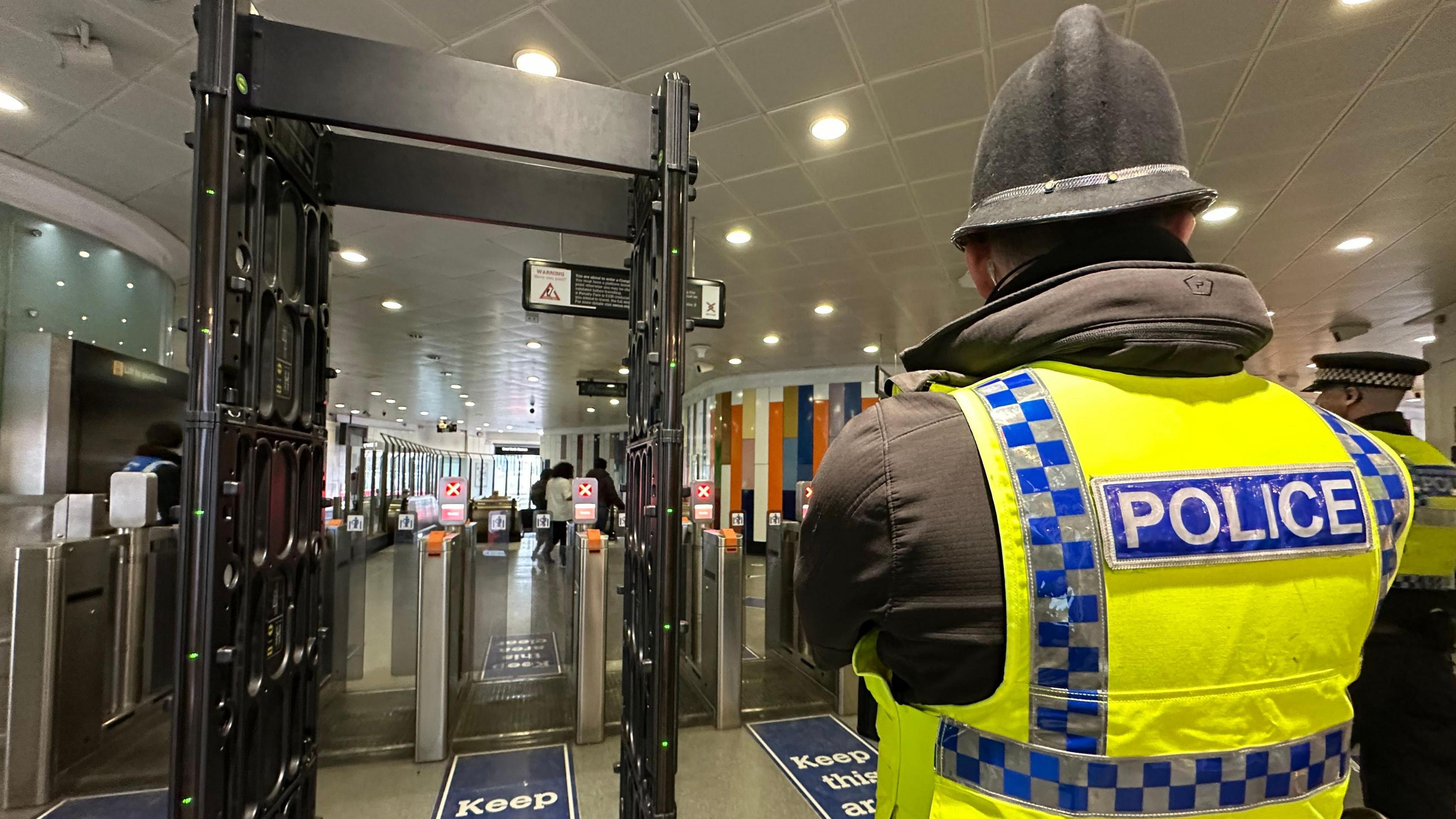 View of the back of a police officer - wearing a hi-vis jacket marked Police, and a helmet - standing to the right of a walk-through metal detector arch placed in front of ticket barriers at Haymarket Metro Station. People can be seen in the background entering the metro station.