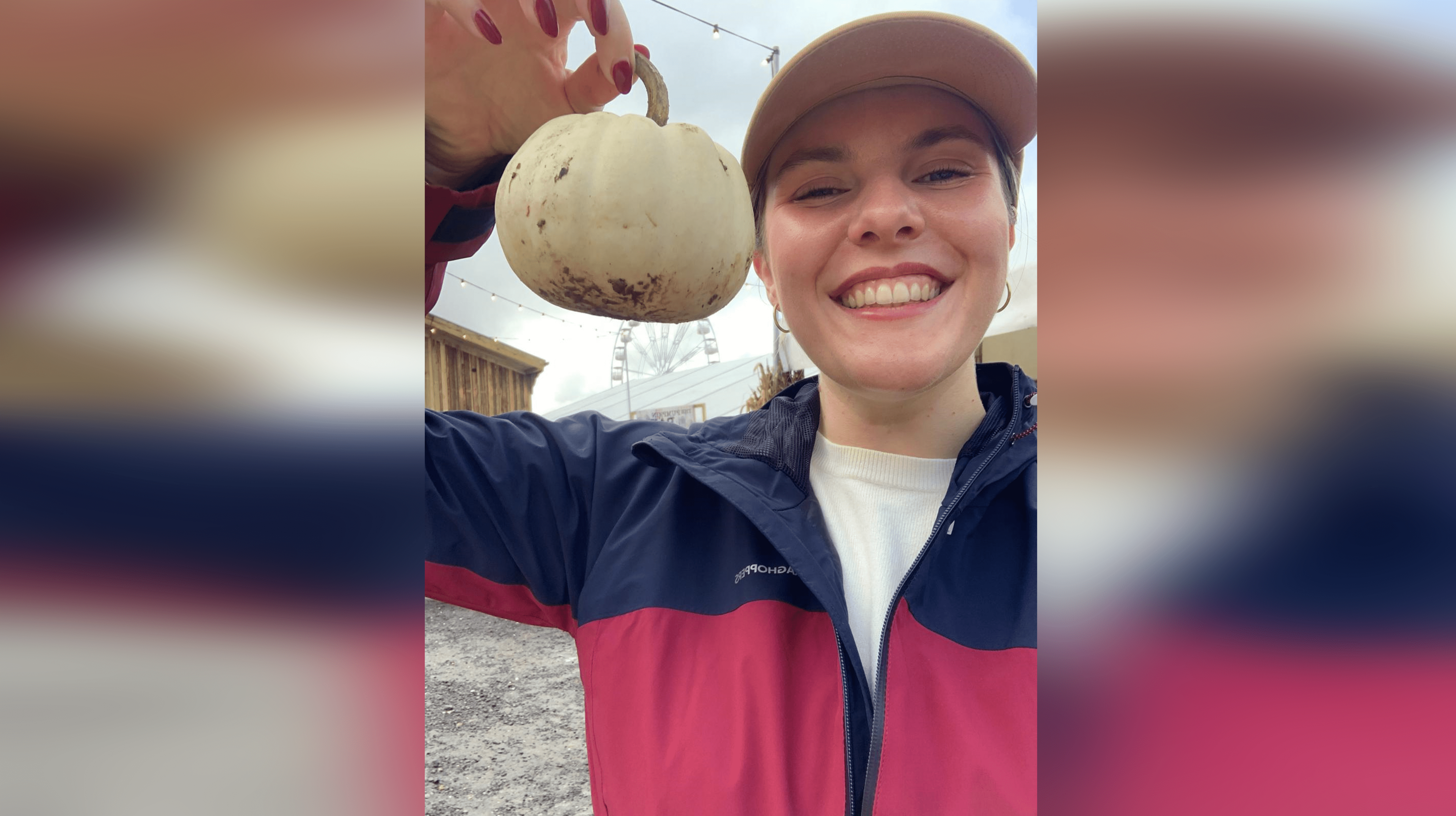 Jess Warren in a blue and burgundy raincoat and beige baseball cap. She holds a white pumpkin next to her head and smiles at the camera.