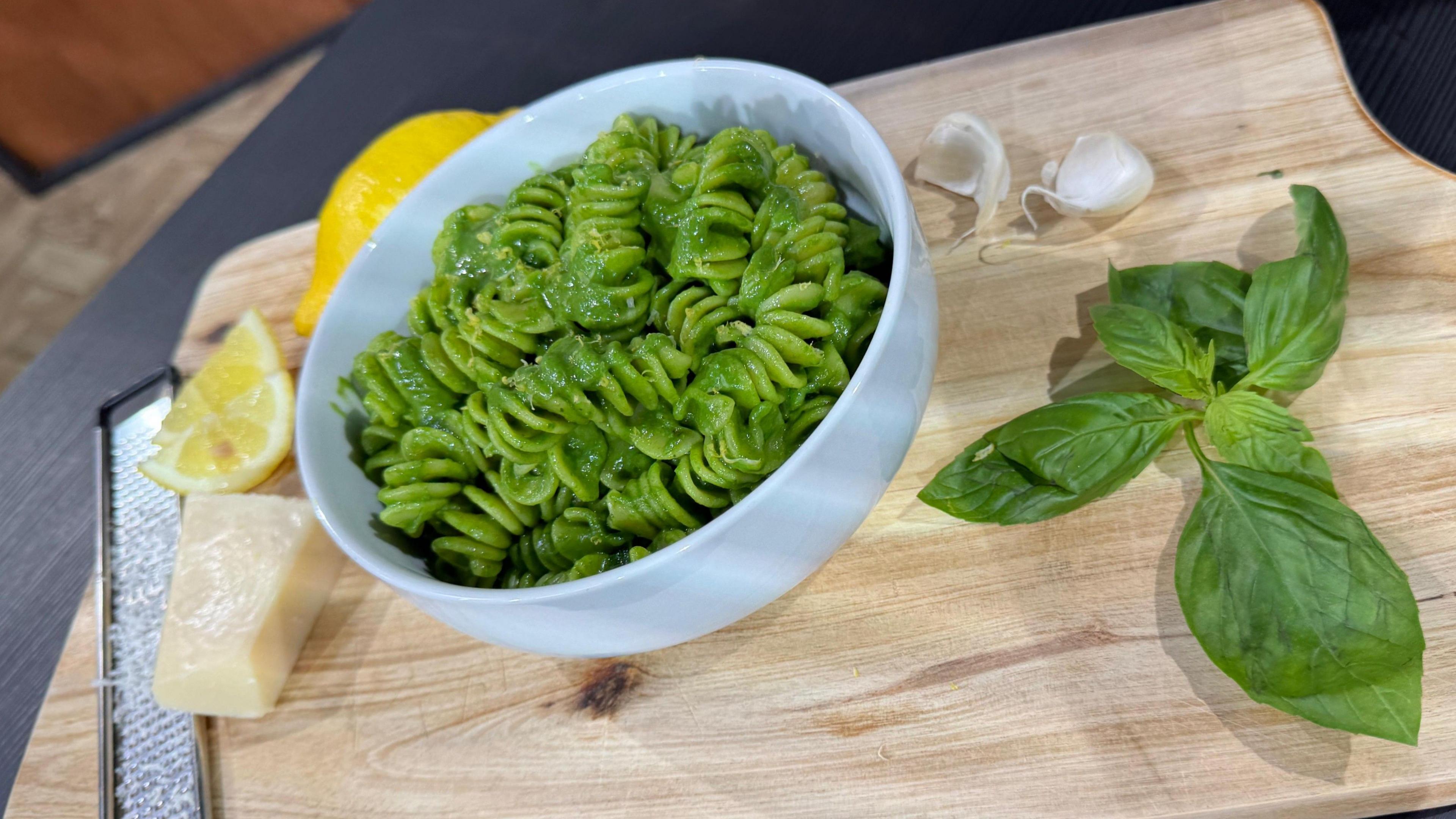 Chris Baber's super green pasta. Twirls of fusili in a white bowl, coated in a deep green sauce. The bowl is on a wooden board and surrounded by basil leaves, cloves of garlic, wedges of lemon and a block of parmesan cheese.