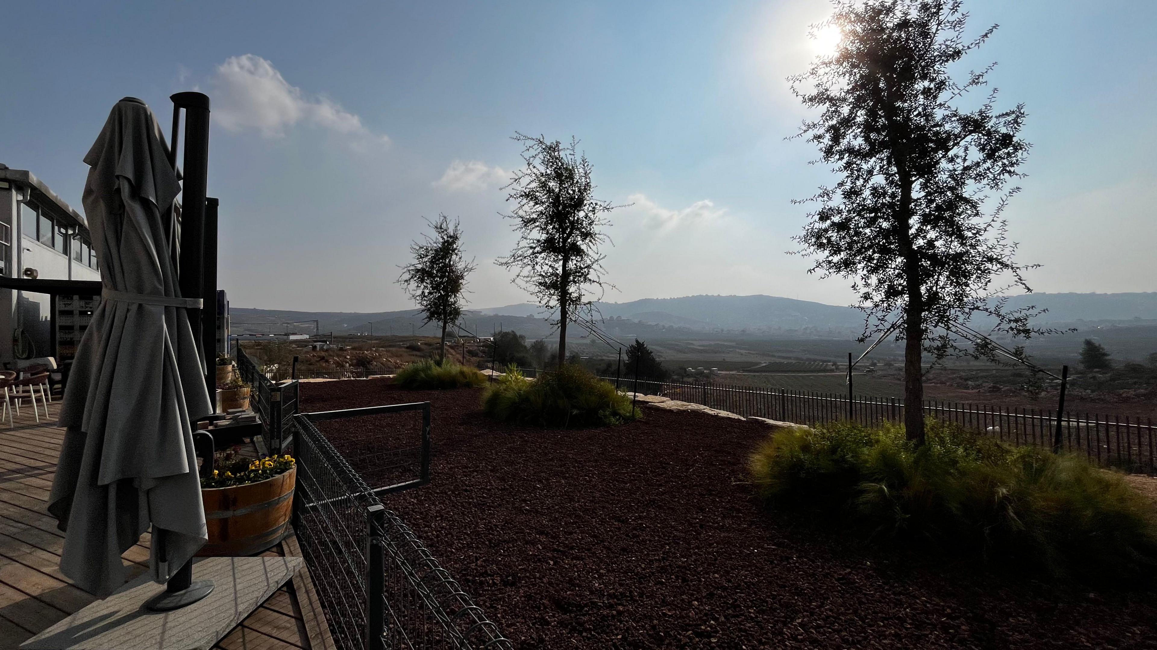 The view from Amichai's winery. There is a deck with an umbrella folded. As you look our from it there are three trees, then a fence then field and hills in the background. There is blue sky with a hint of cloud and the sunlight gives the image a misty feel