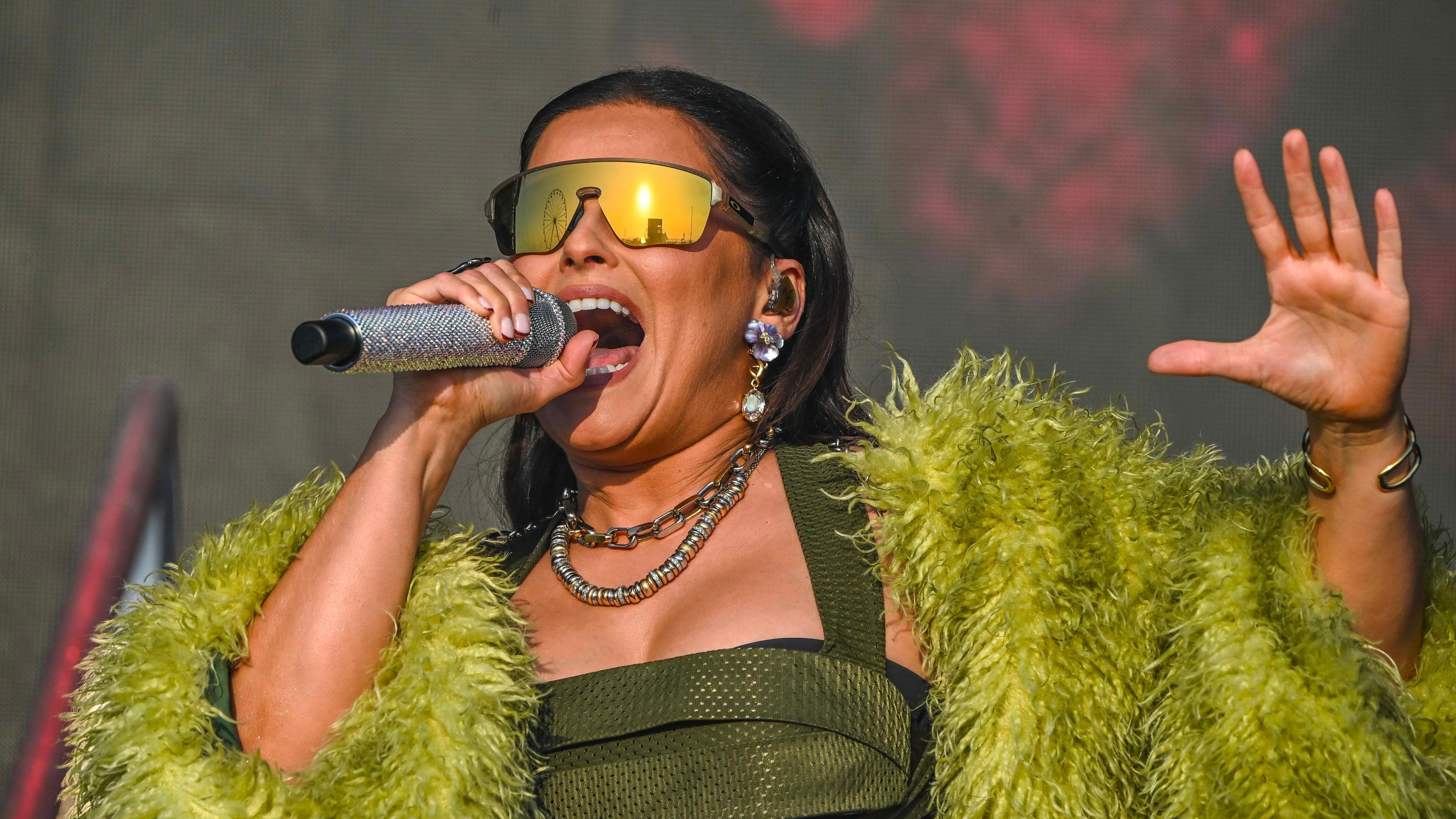 The image shows a person holding a microphone with a glittery handle, dressed in a green, fluffy-textured outfit. They are wearing large earrings and multiple necklaces, with a dark background featuring red lighting effects.
