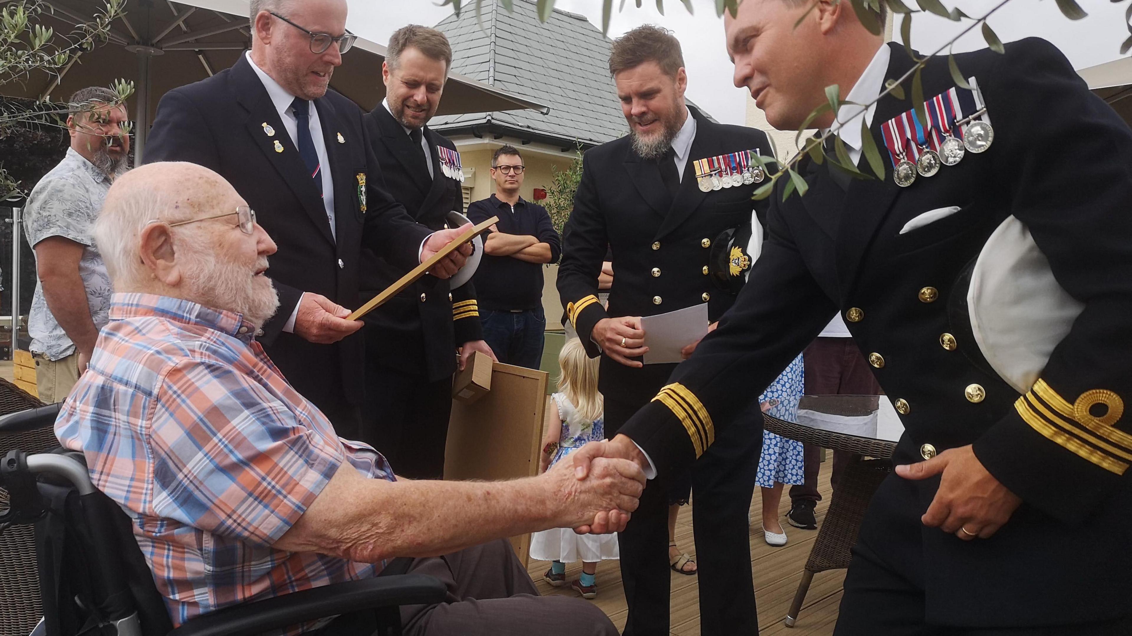 An elderly man in a check shirt sitting in a wheelchair and shaking hands with a man in a naval uniform wearing a number of medals. In the background there are a number of other men in naval uniforms along with civilians and children