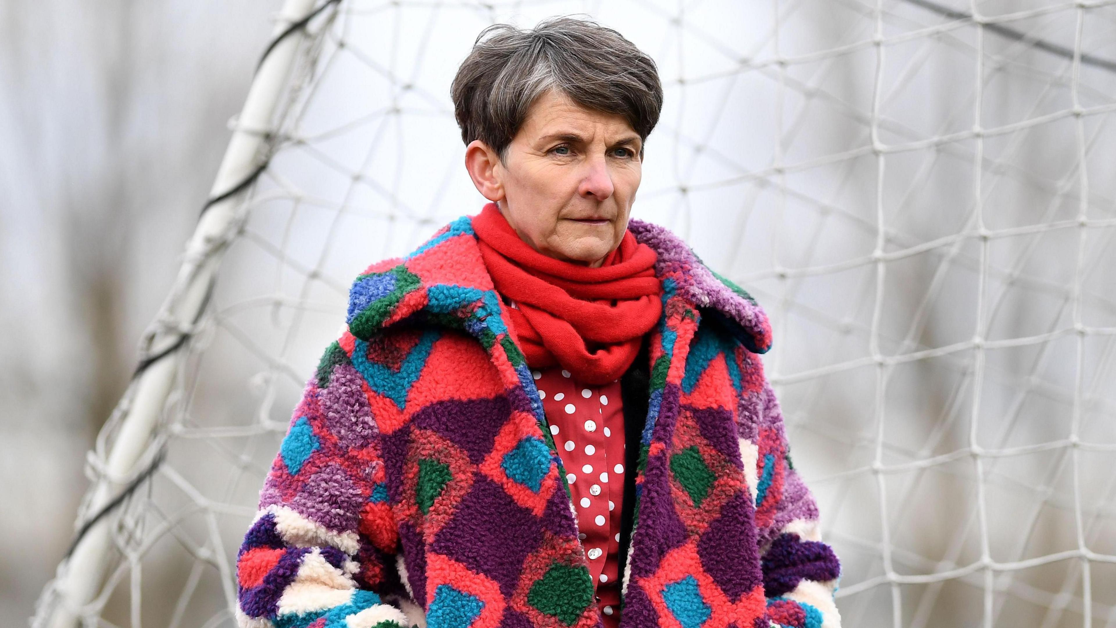 Laura McAllister is standing in front of a football goal. She is looking to the right of the image. She is wearing a red scarf and a red and white polka dot top. She had a woolly coat over the top which is in the colours of purple, blue, green, pink and white. She has grey hair.