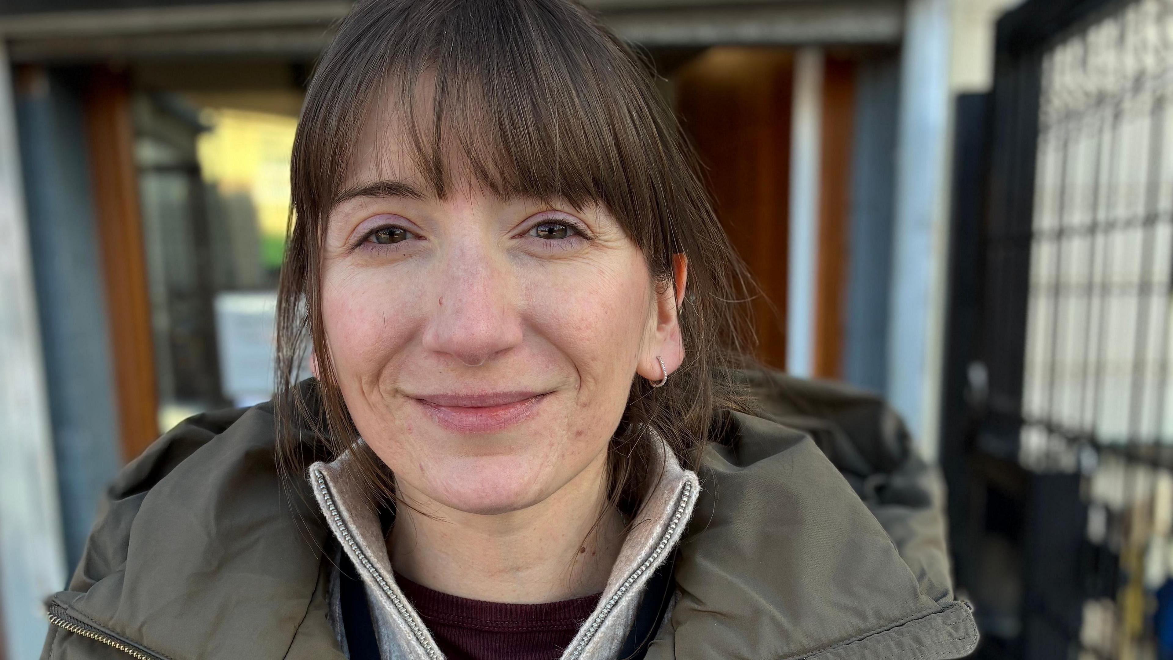 Lucy Cox stands outside a building. She is wearing a jumper and winter jacket and has her brown hair tied back.