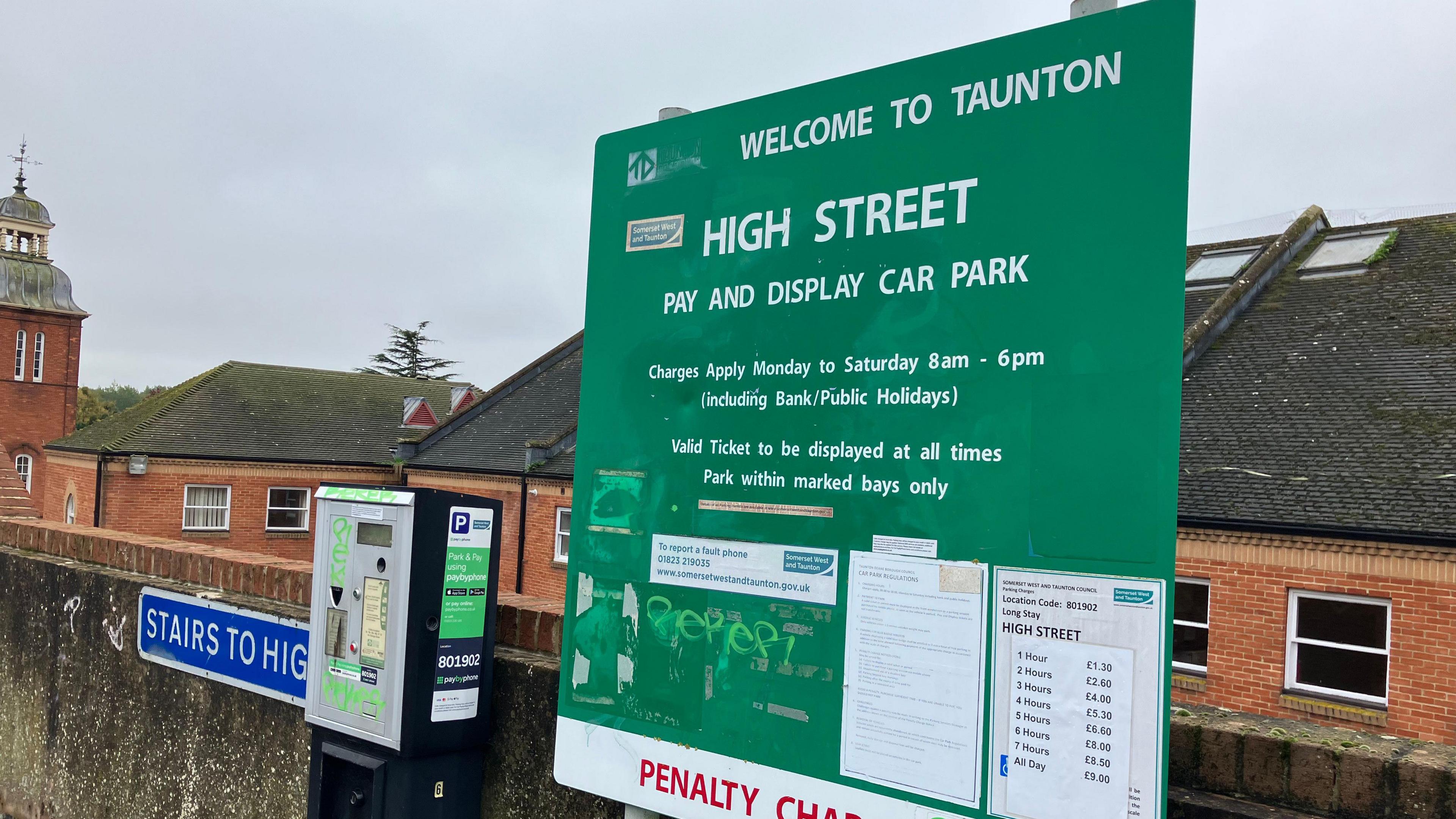 The green pricing board for the car park, saying 'Welcome to Taunton'. There is some graffiti on the board. On its left is the parking meter, next to the a sign saying 'stairs to high street. In the background are brick buildings and a grey sky.