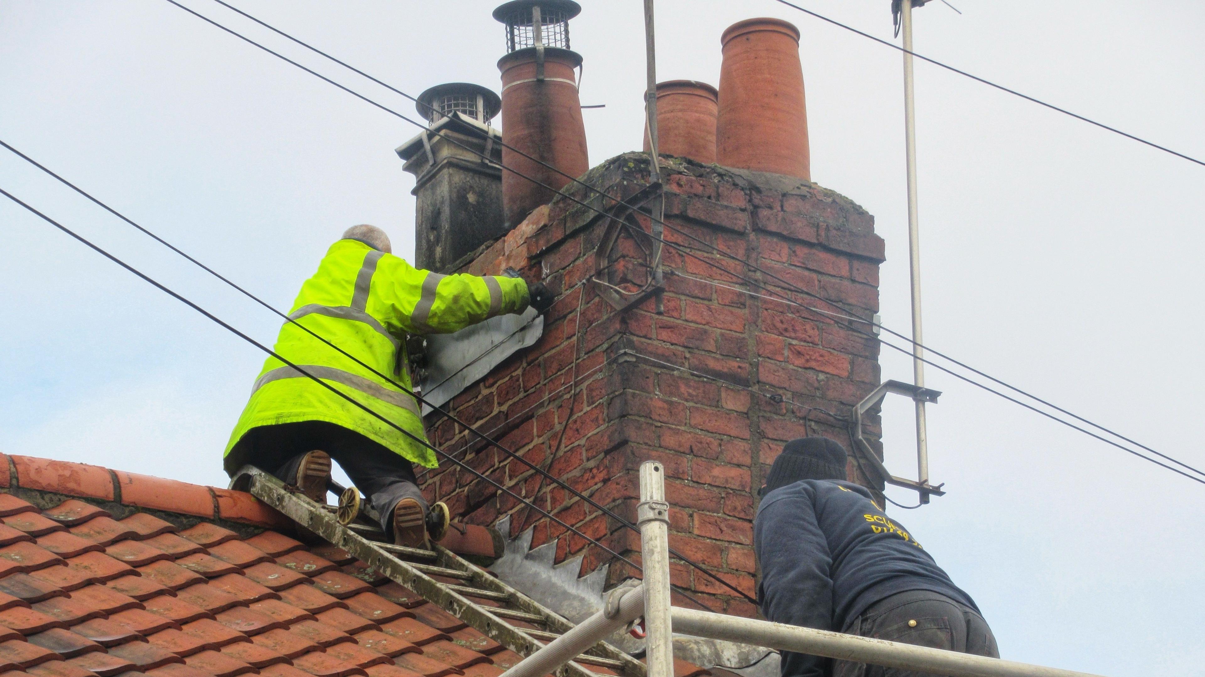 Two men, one wearing a hi-vis jacket, are on a roof. One is fixing a panel to the chimney. Scaffolding has been put up around the house and there is a ladder laid over the roof.