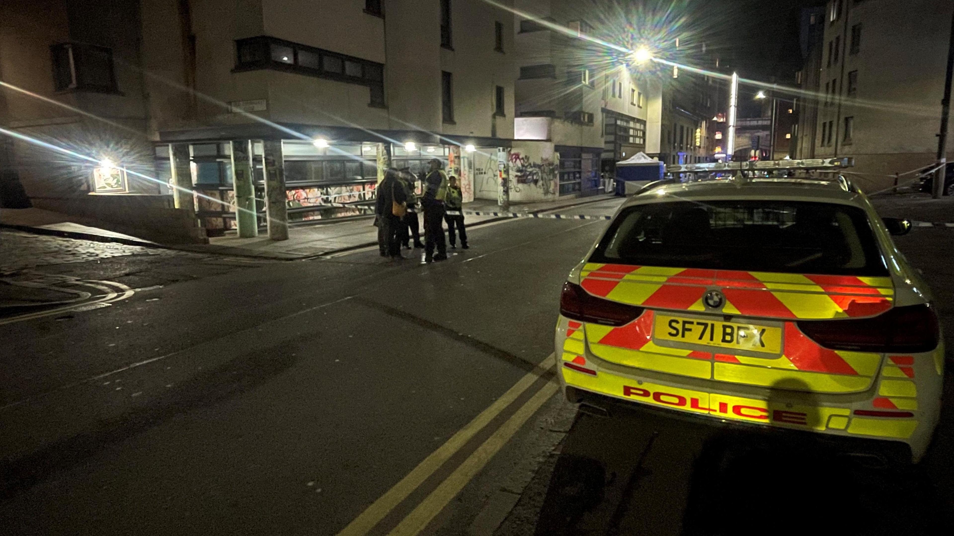 A police car parked on the pavement in the Cowgate in Edinburgh. There is a double yellow line on the road. A police officer stands in front of the fence talking to several people in dark clothing.