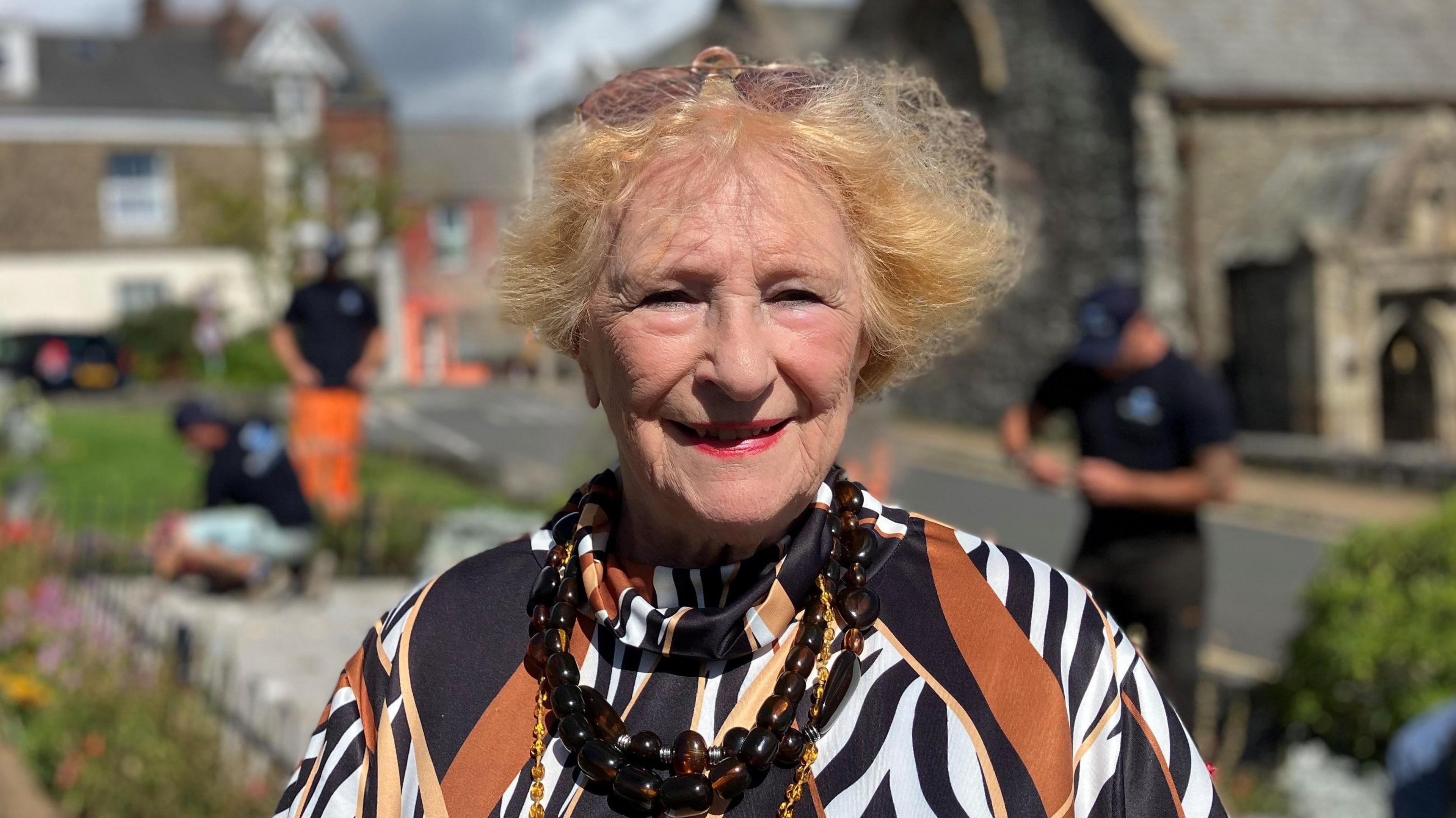 Older lady with red hair smiling at the camera wearing big beads and a silk blouse of tiger print