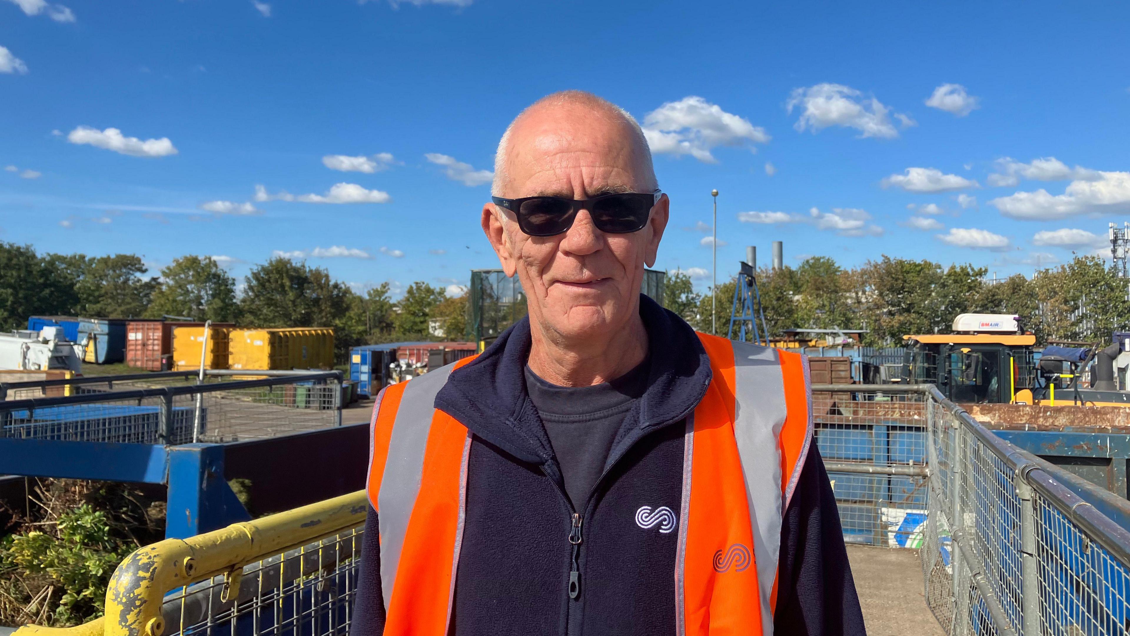 Gary Skinner looks at the camera and smiles. He is wearing sunglasses and has a high-viz orange waistcoat over a dark blue fleece. Behind him is the tip site, with metal fences and containers throughout.