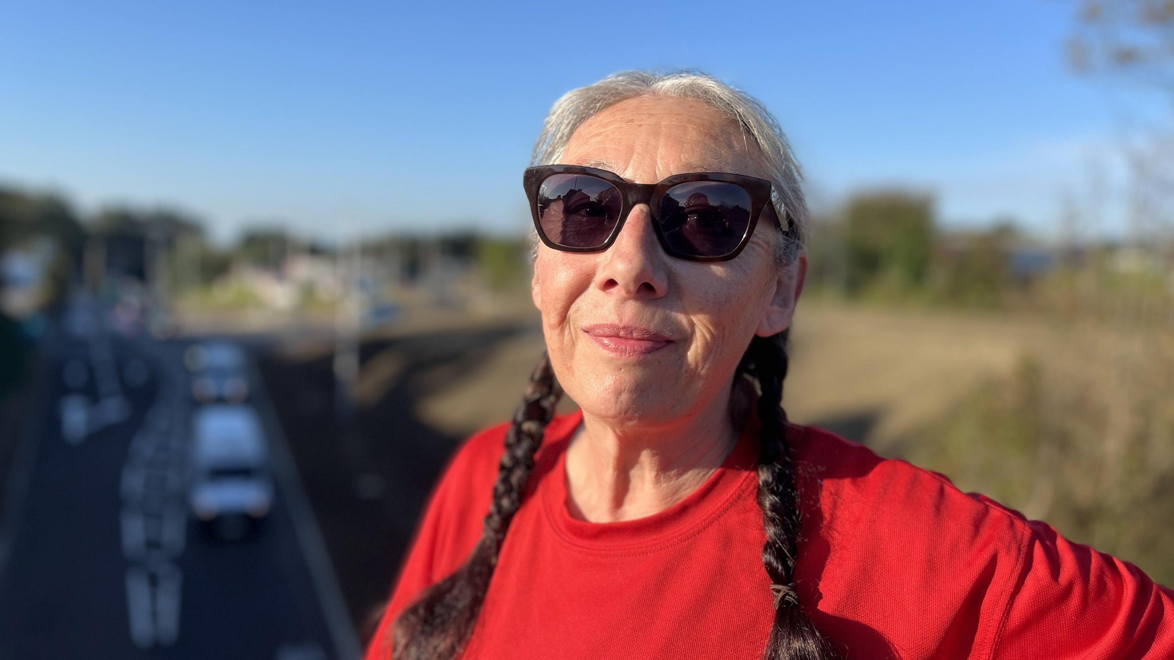 A woman with plaits in her hair and sunglasses wearing a red t-shirt. There is a road out of focus in the background.