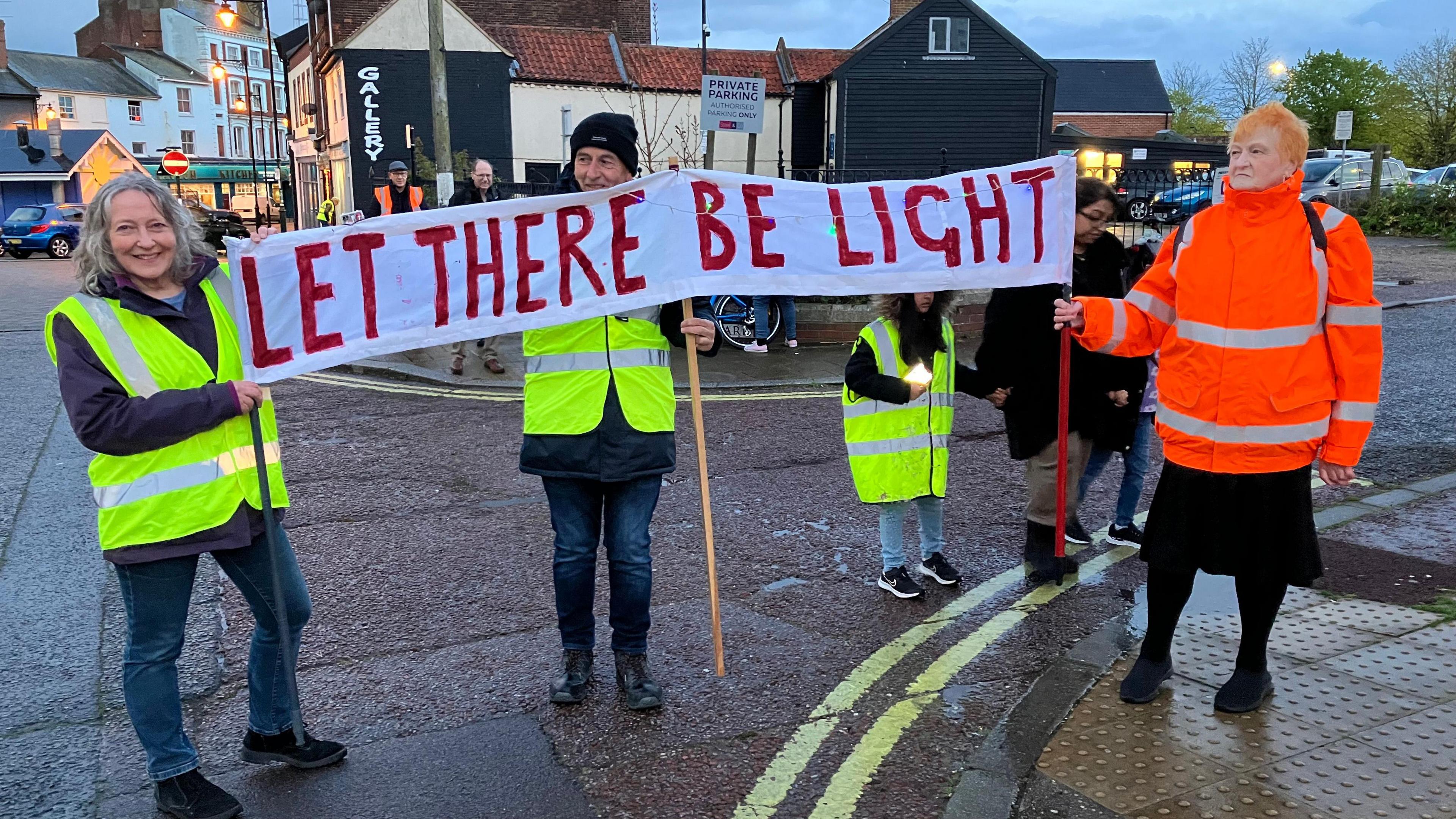 Campaigners wearing yellow and orange fluorescent jackets hold up a sign reading "Let There Be Light" while on a street protest march