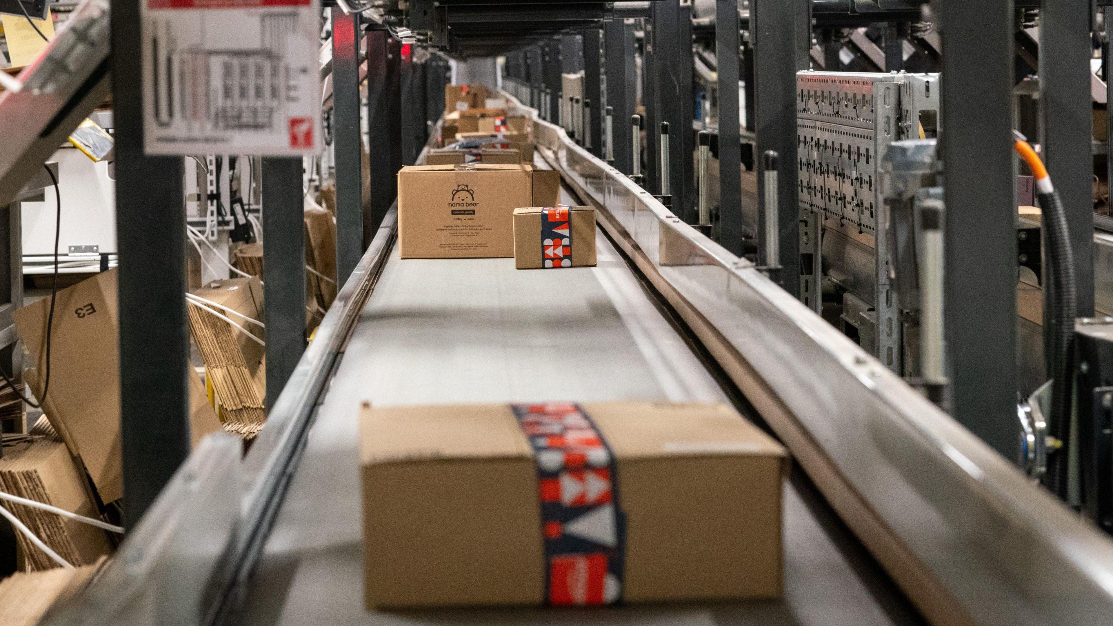 Brown boxes with red, white and navy coloured tape wrapped around them make their way down a conveyor built.