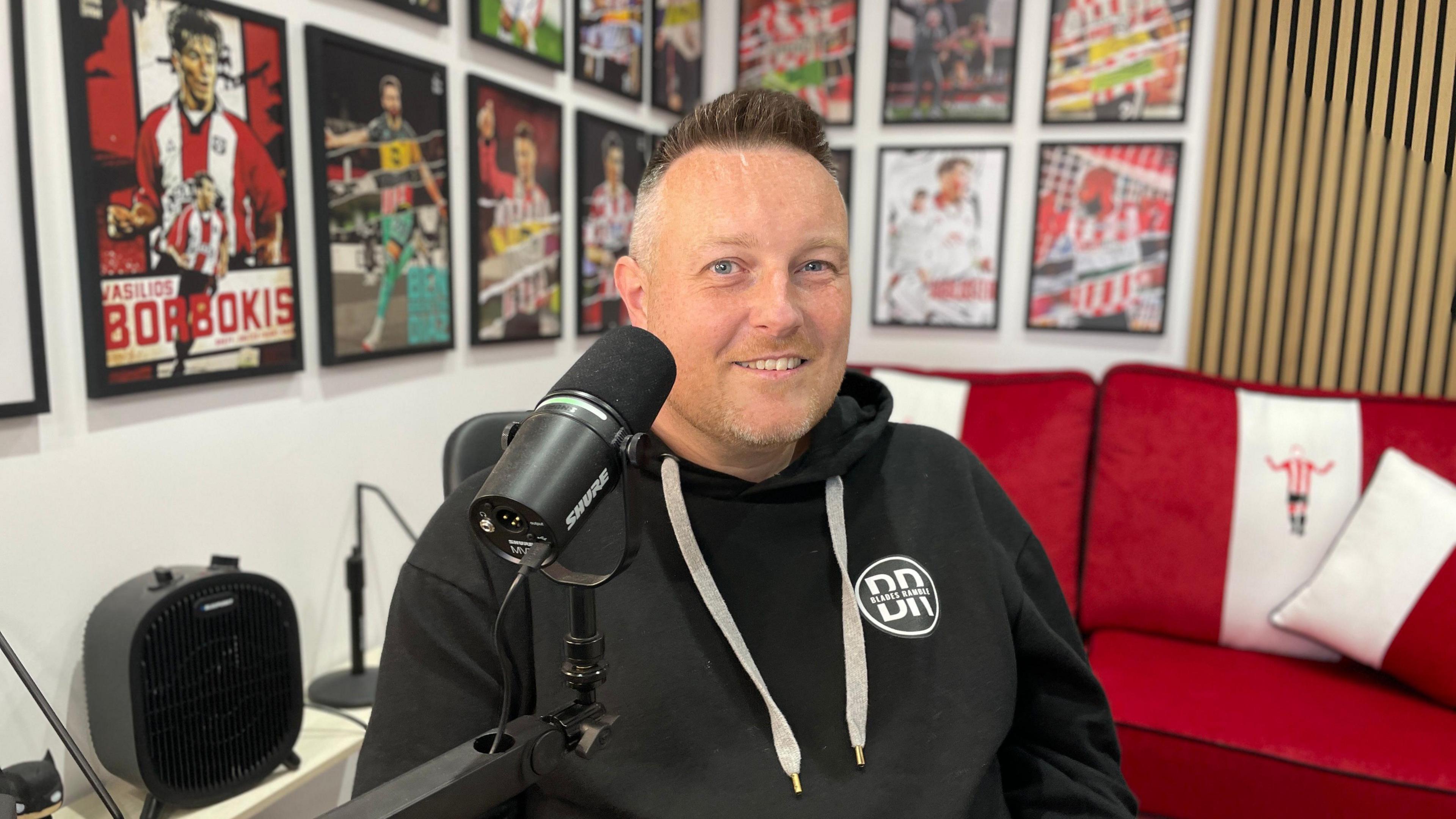 A picture of a smiling Jimmy Smith. He has short hair and is wearing a black hoodie. He is sitting next to a microphone. Behind him is a red sofa and a wall filled with framed Sheffield United posters.