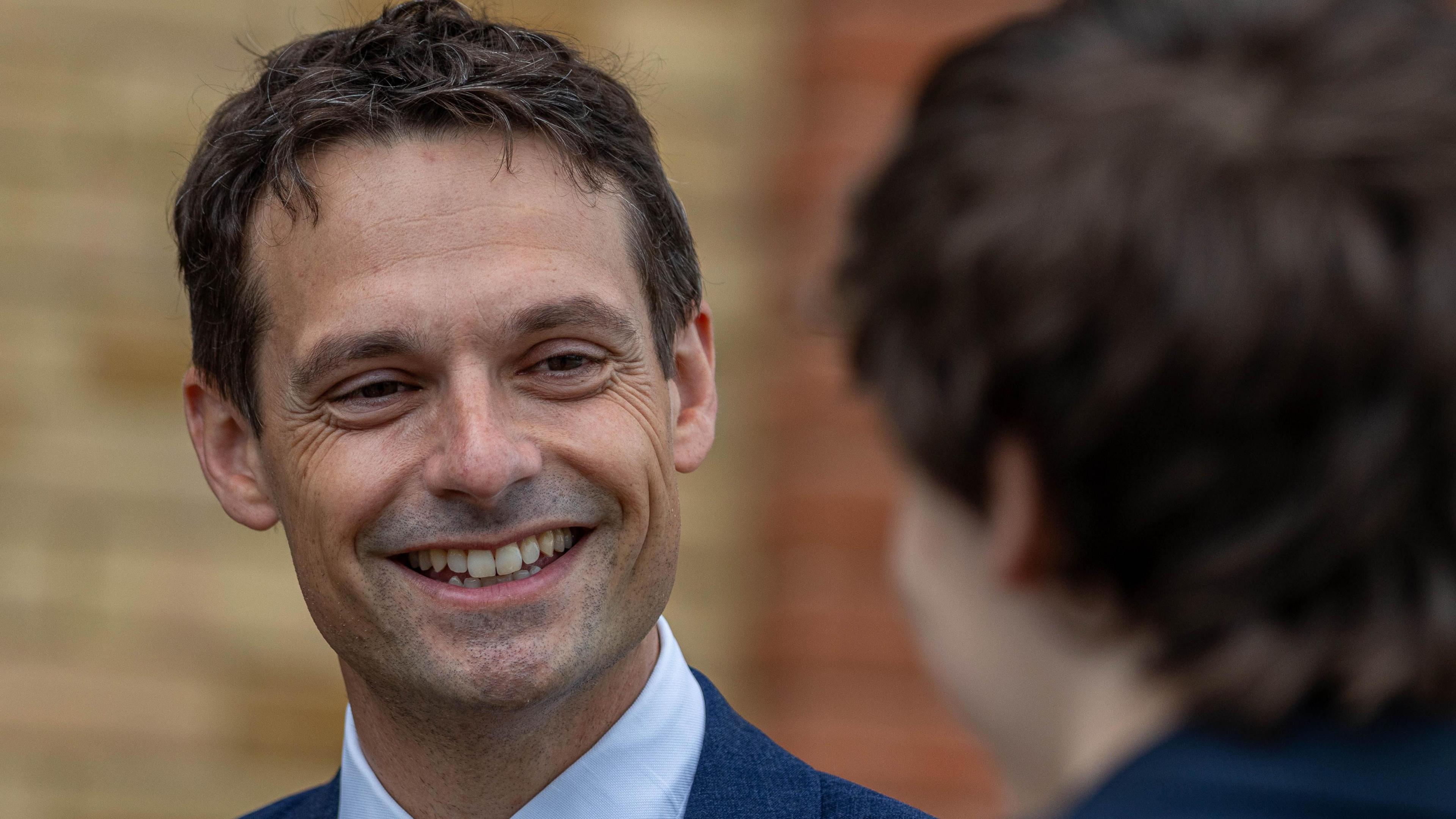 Image shows Pate's Grammer School's head teacher James Richardson. He is smiling and looking at a student. The student has their back to the camera and is out of focus. Mr Richardson has brown hair and is wearing a white shirt and blue blazer. 