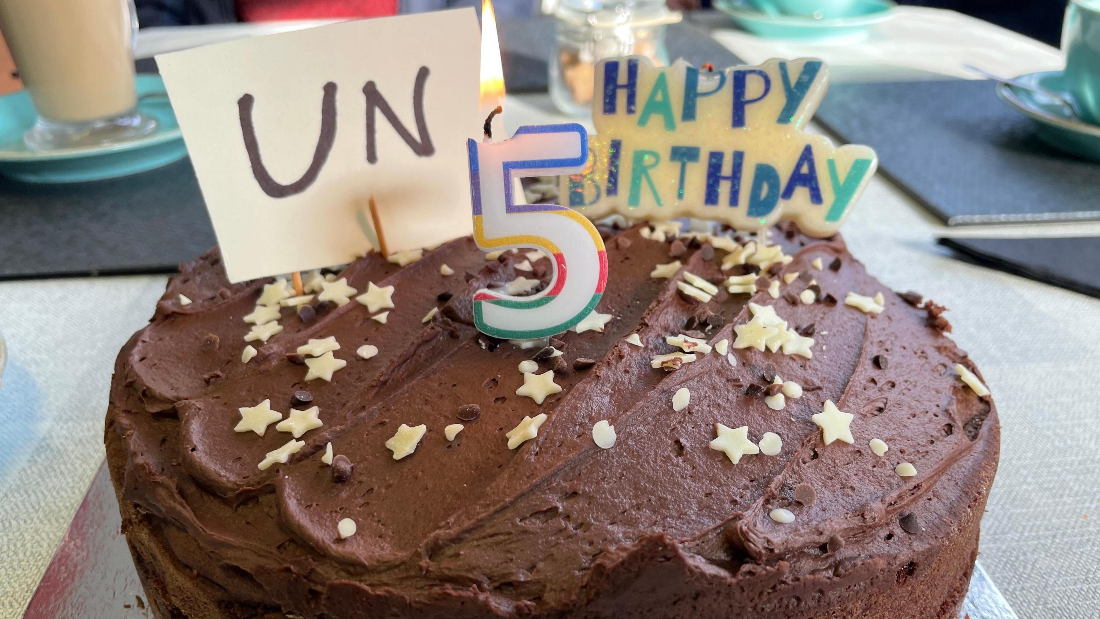 A close up image of a birthday cake. It is a chocolate cake with cream coloured stars and circles sprinkles on top. There is a candle in the shape of the figure five and another candle says "happy birthday". Next to it there is a piece of paper stuck into the cake that reads un, to spell out unhappy birthday in full.