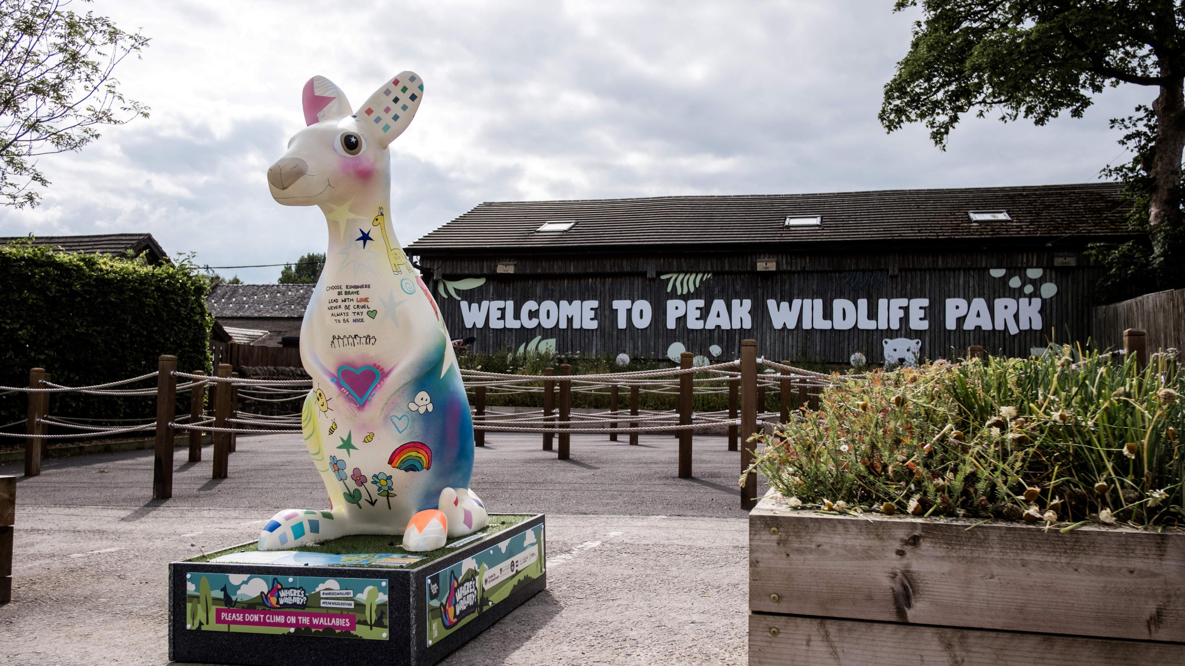 A sculpture of a wallaby, situated at the entrance of a wildlife park. It is colourfully decorated and there is a raised flowerbed next to it.