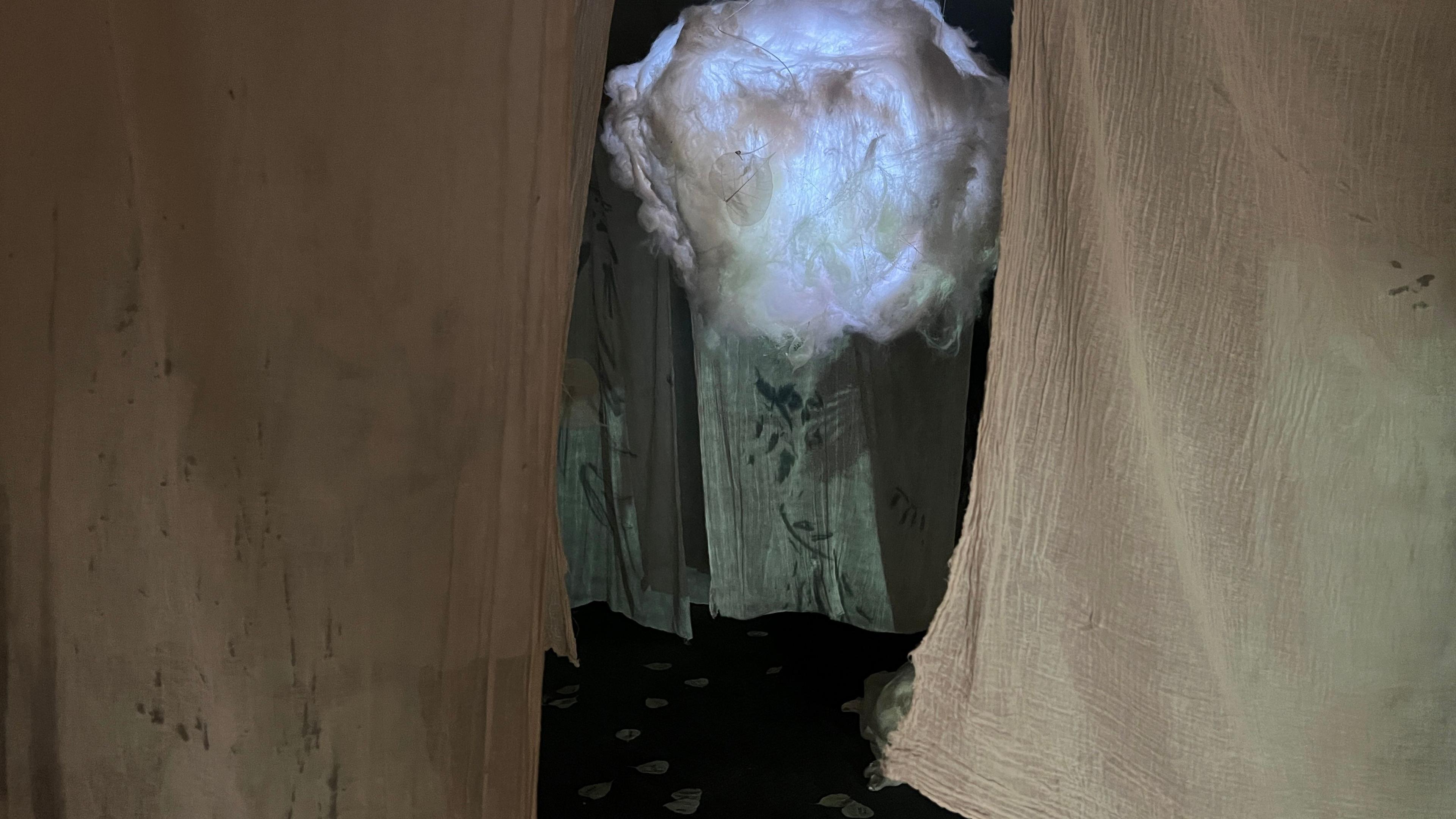 Sheets are hanging from the ceiling in a small room. There is a construction made to look like a cloud in the middle, which is white and lit up.