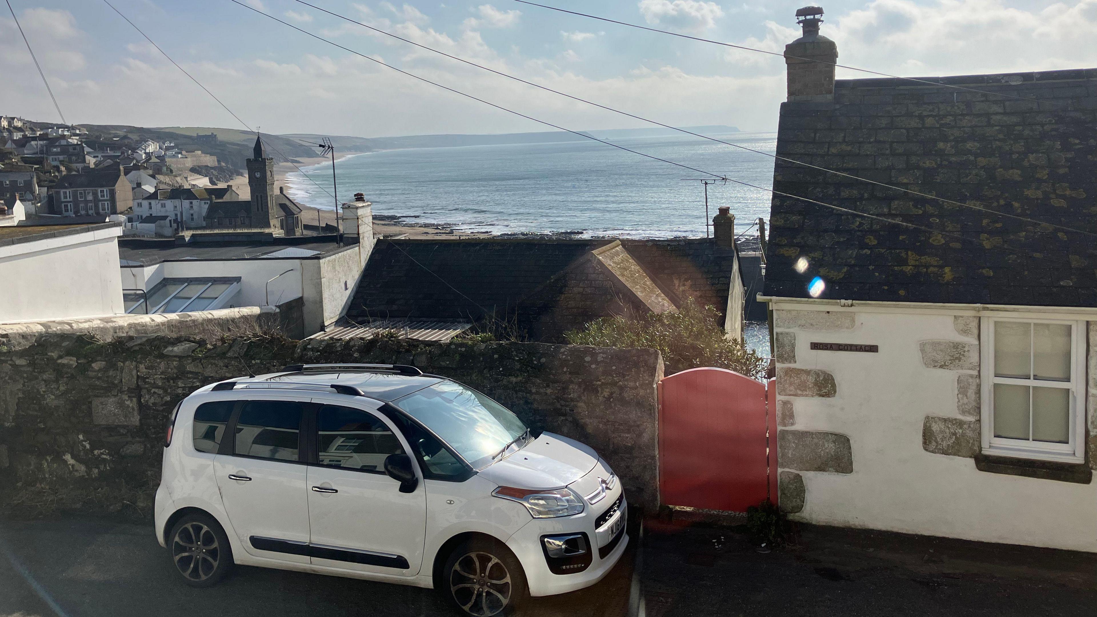 View of Porthleven and over the sea. In the foreground is a parked car on a lane and part of a single storey house.