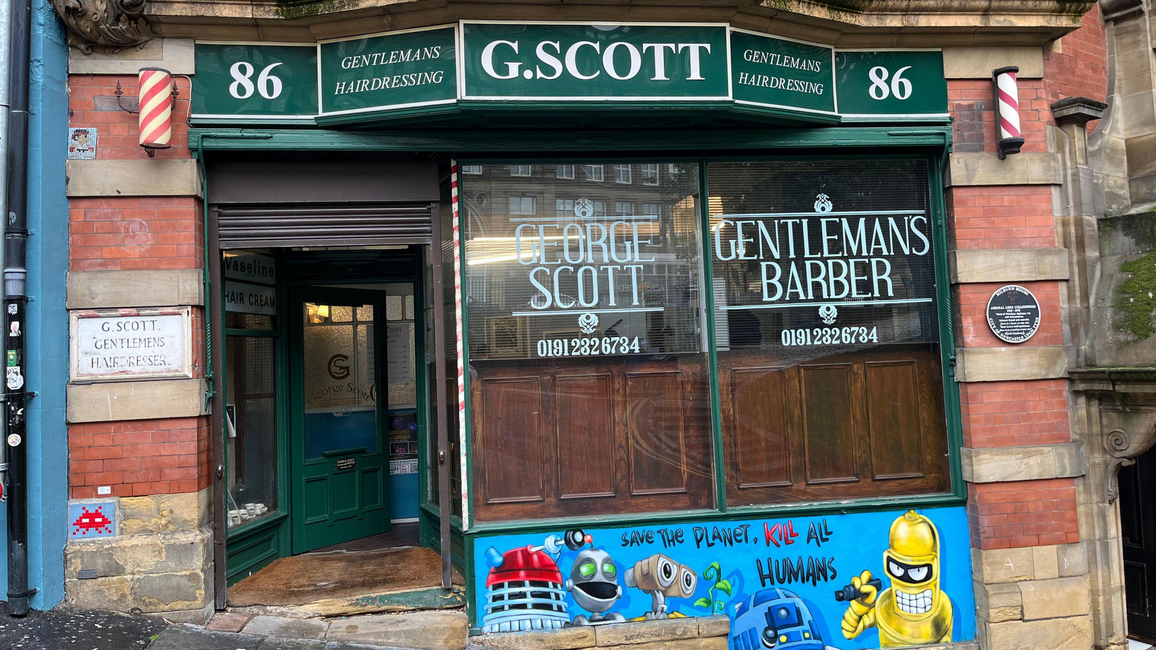 The outside of George Scott Gentleman's Hairdressing. The shop sign is green with white lettering. Two small, red and white barbers poles hand on either side of the sign. An older sign carved into the stone also states the shop's name. Under the shop window there is a more modern painting of robots from popular culture, such as R2D2 and a darlek.