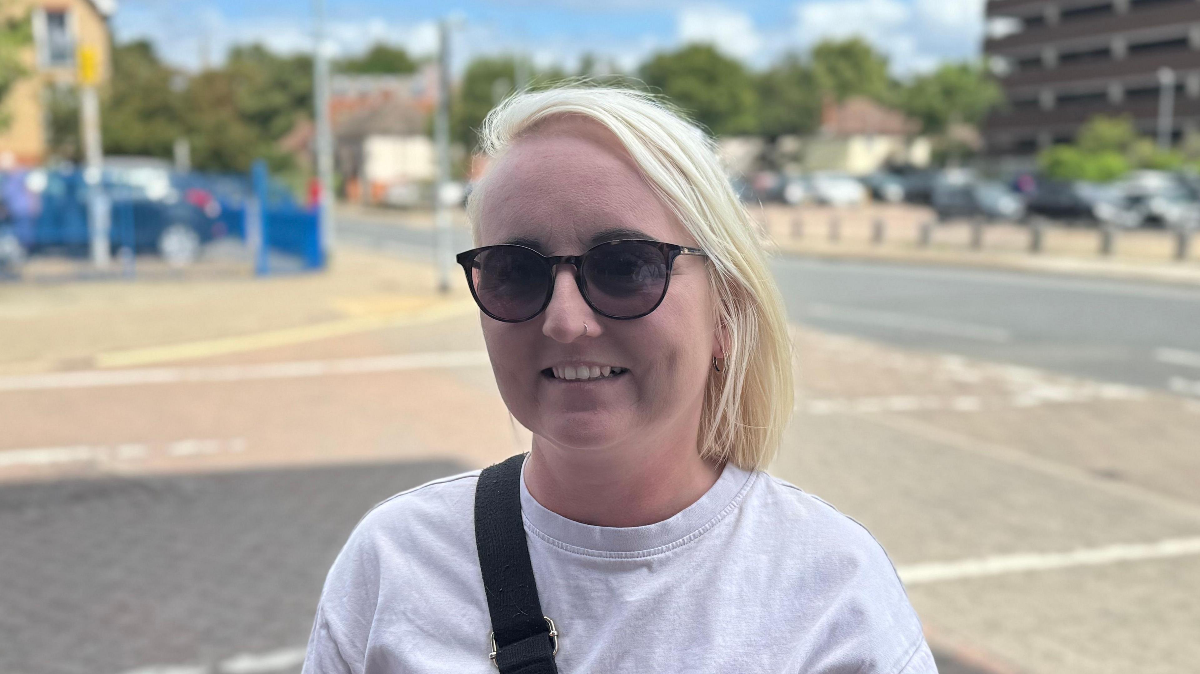 A head and shoulders picture of a woman with blonde hair. She is wearing a light T-shirt and sunglasses. She is smiling while looking into the camera.