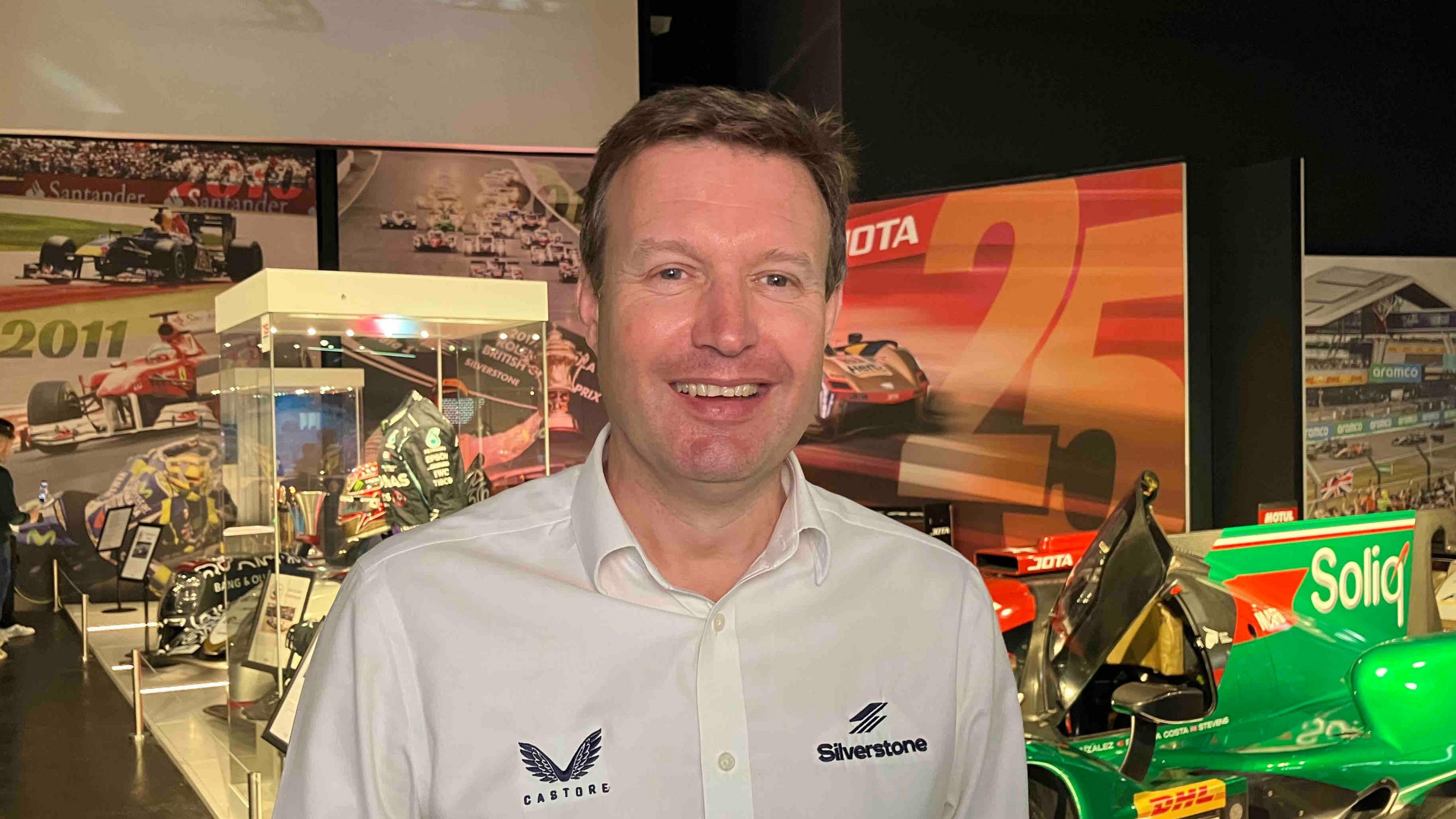 Stuart Pringle with short brown hair smiling at the camera and wearing a white T-shirt with the Silverstone Logo. He is standing in front of display cases at an exhibition.