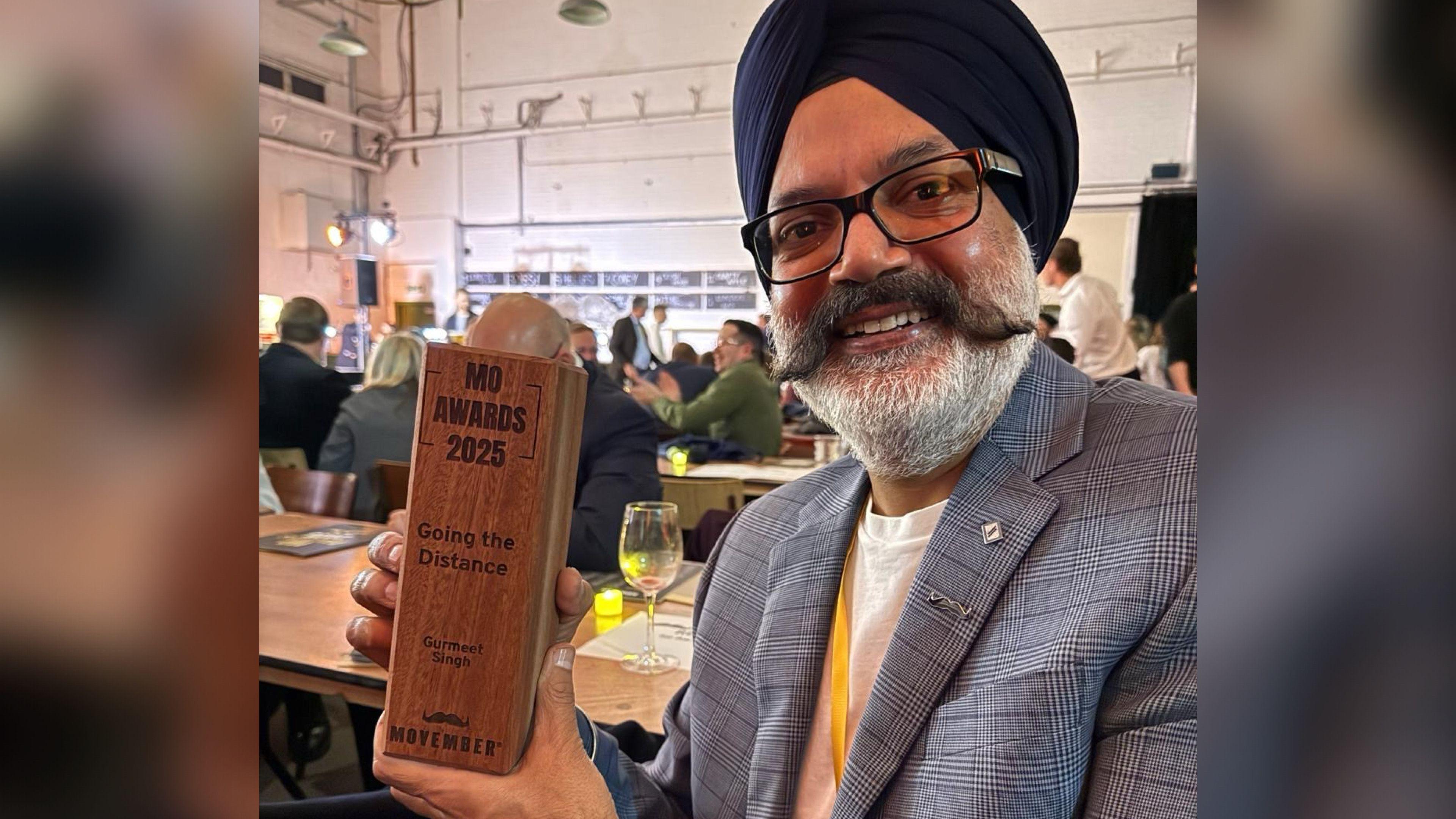 A man with a white beard and dark moustache wearing a turban, glasses, white shirt and grey patterned suit smiling at the camera and holding a wooden award that reads: "MO AWARDS 2025. Going the Distance. Gurmeet Singh. Movember".