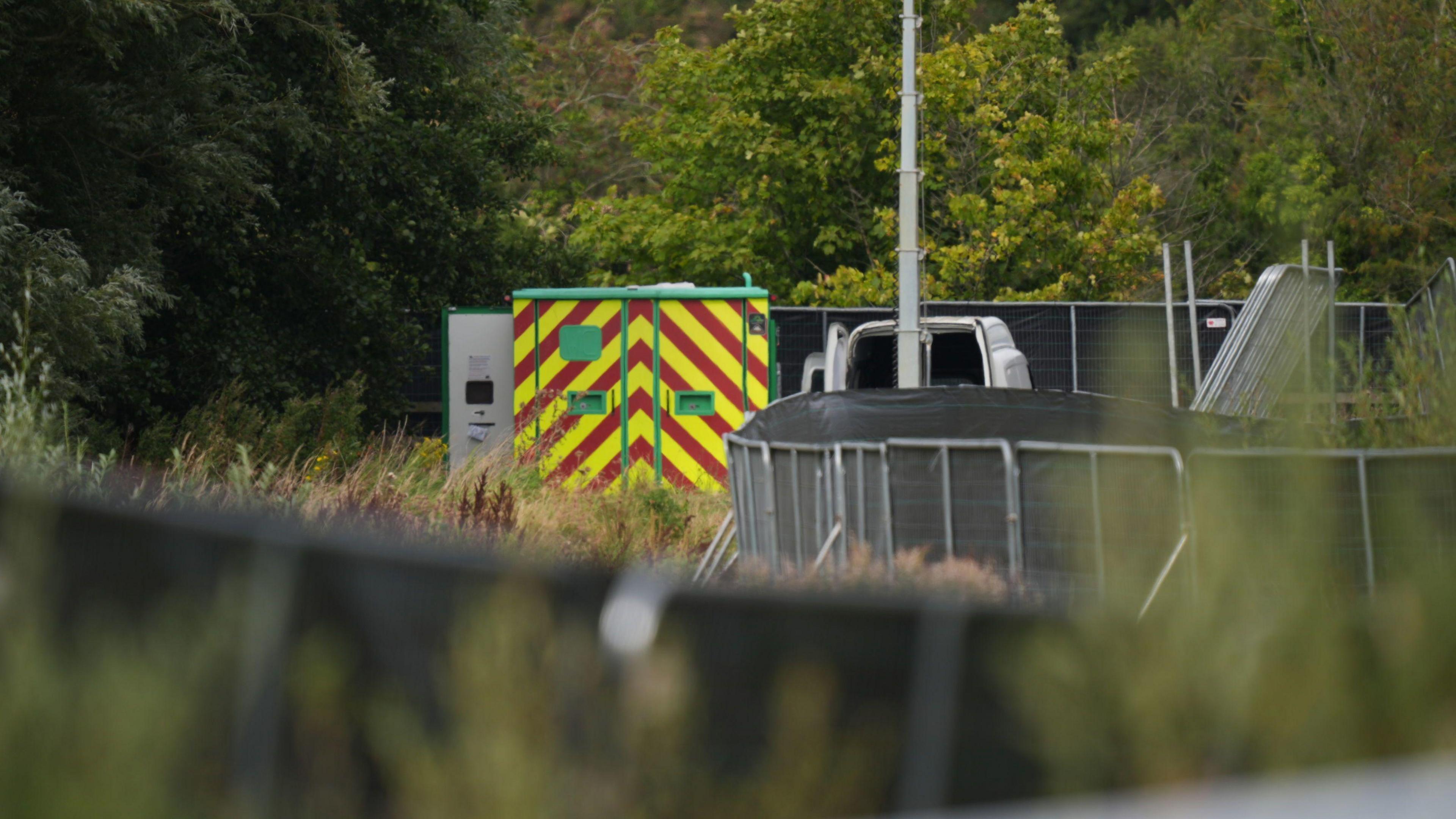 A large red and yellow tripe container sits behind a metal fence with balck covers. The container is surrounded by green woodland.