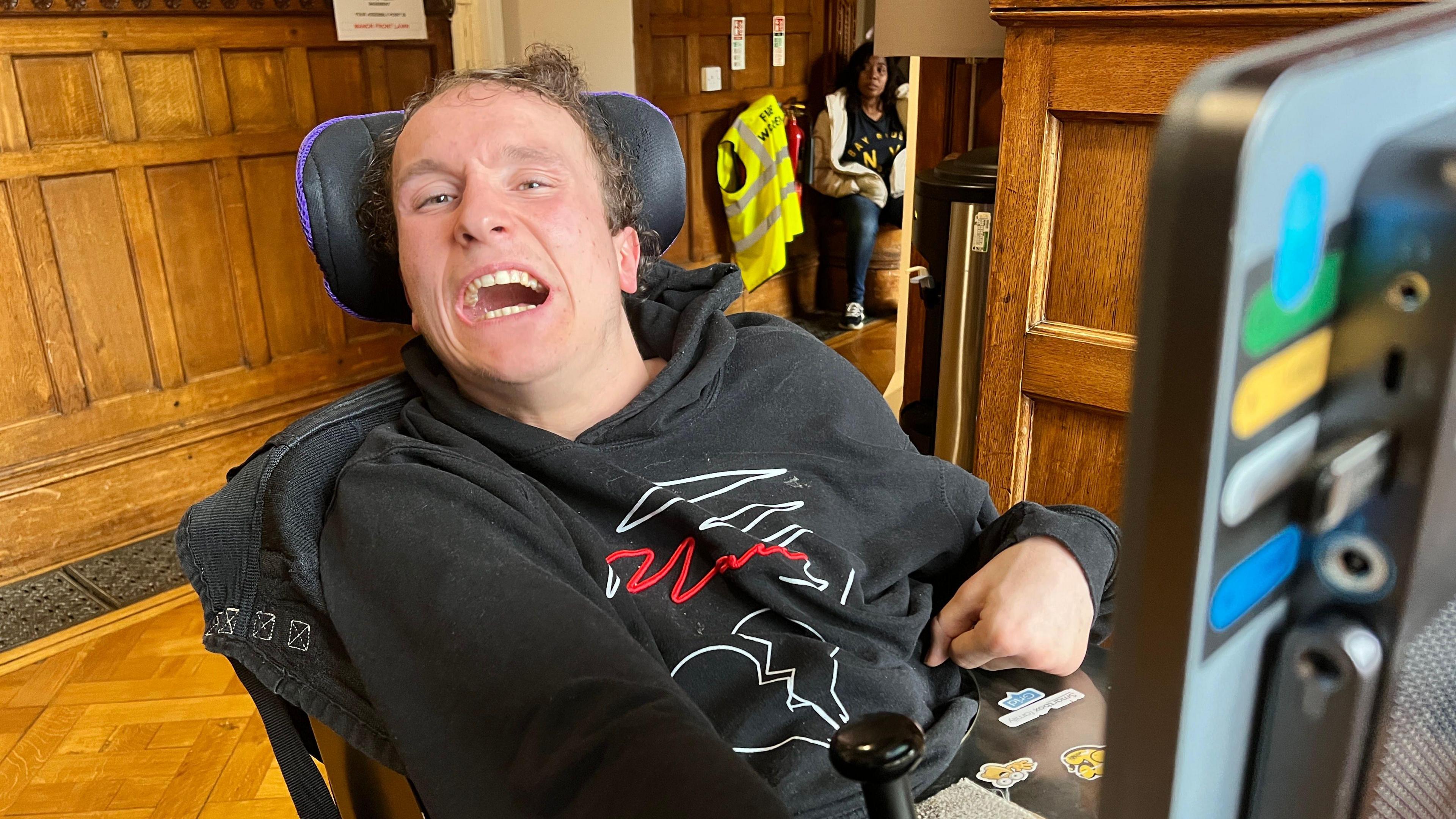 A person seated in a powered wheelchair wearing a black hoodie with a white and red graphic design. A mounted communication device is visible in the foreground. The setting includes wooden panelled walls, a parquet floor, and a doorway in the background.