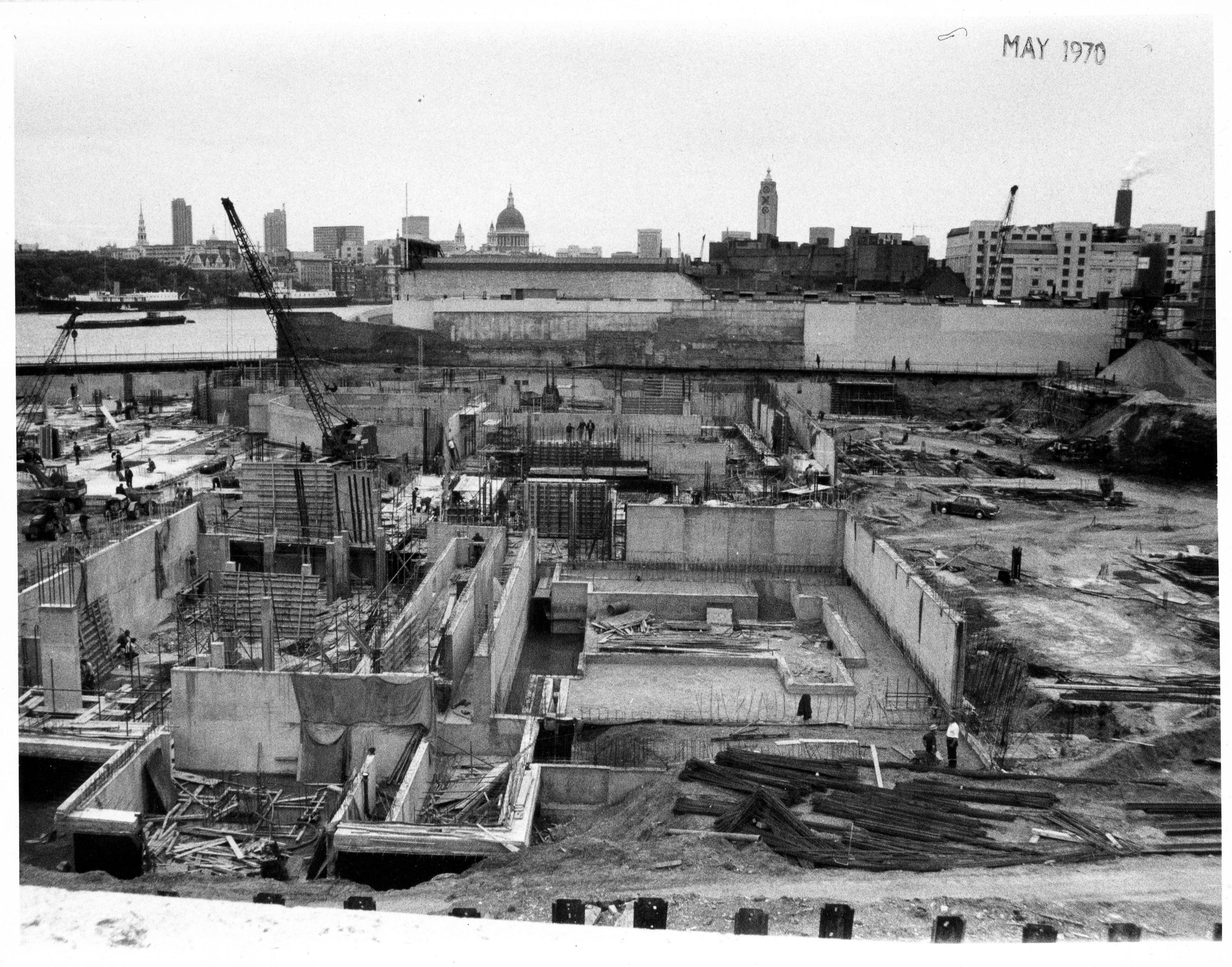 Builders working on the lower concrete terraces of the National Theatre while it was under construction. In the distance, other buildings can be seen including St Paul's Cathedral. 
