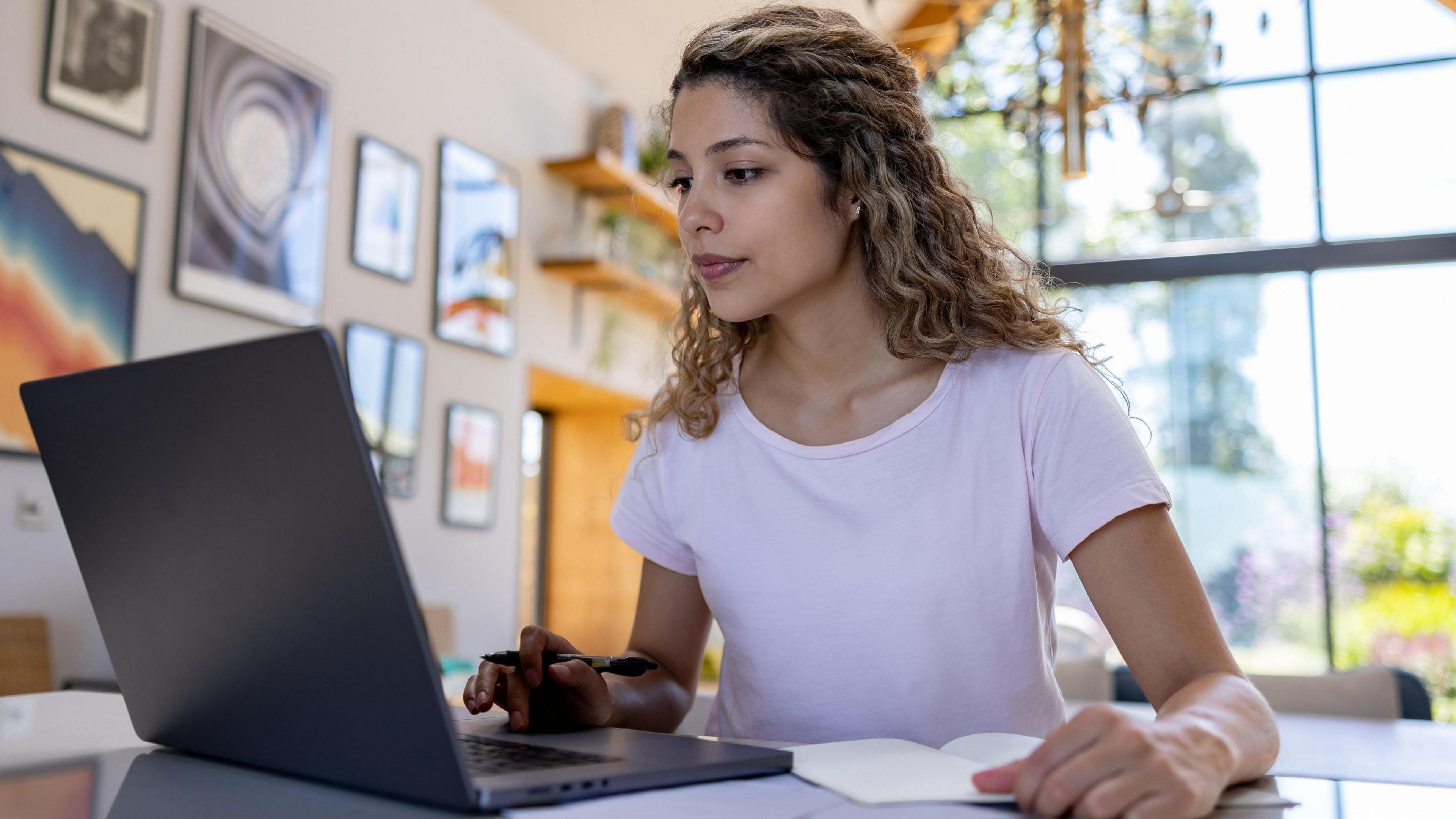 A woman looking in to a laptop with a pen and paper nearby