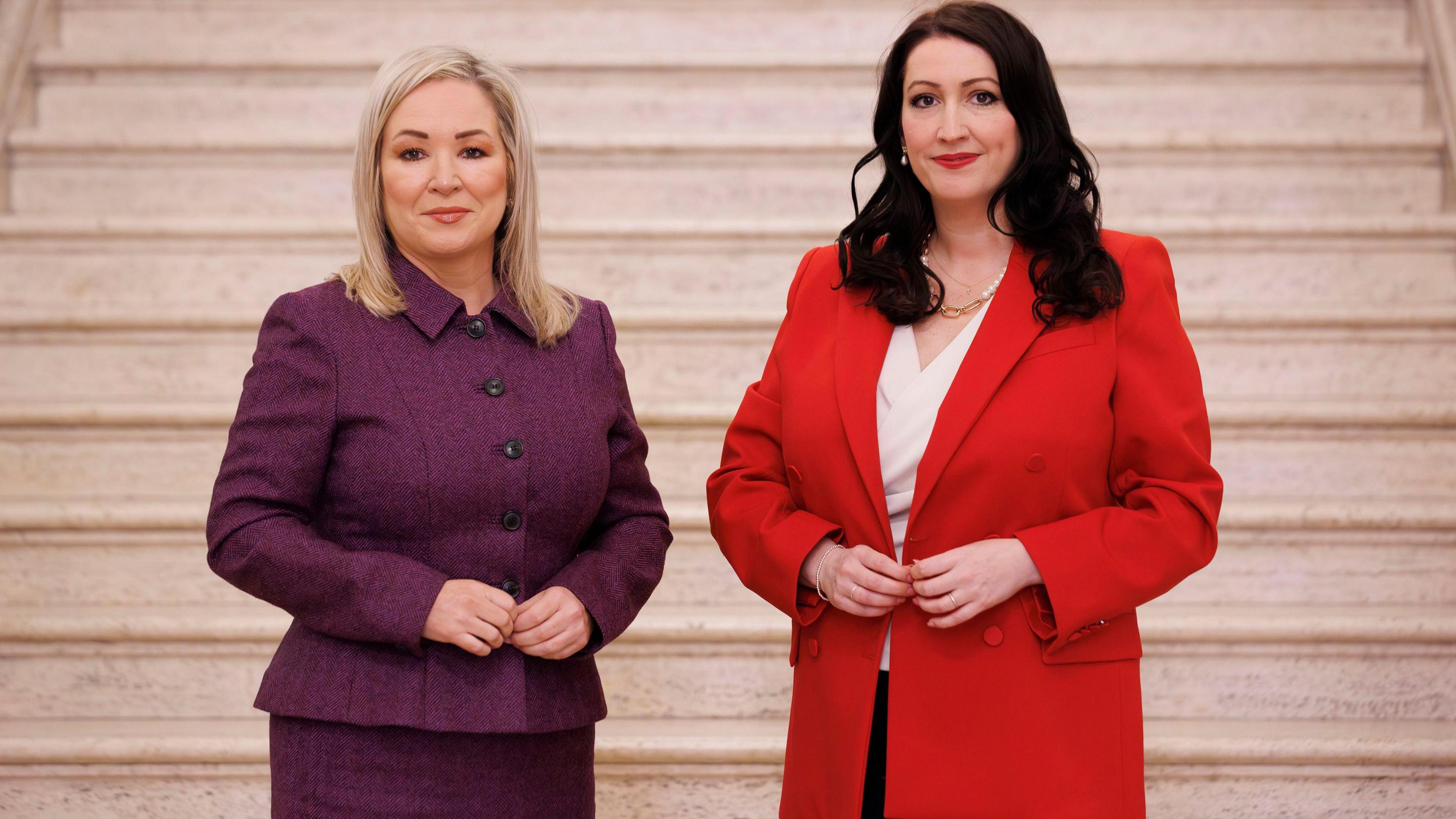 Michelle O'Neill wearing a purple suit, she has blonde hair. Emma Little-Pengelly in a red blazer and white top, she has black hair.