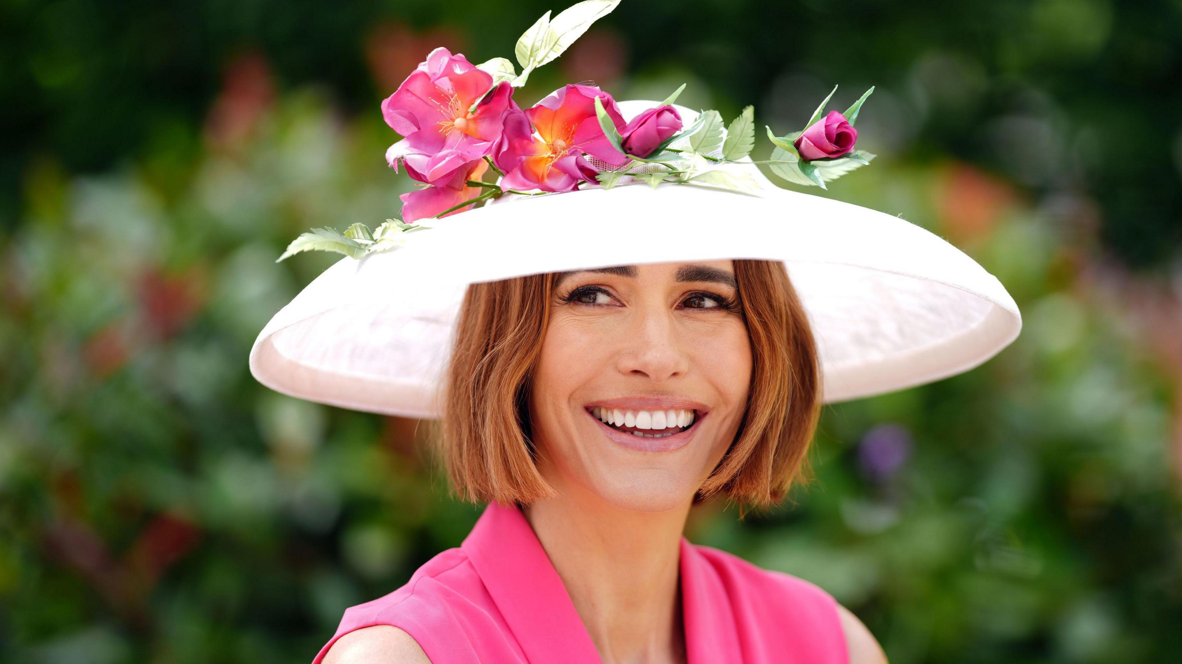 Louise Roe, a white middle aged woman with light brown short hair smiles wearing a wide brimmed white hat decorated with pink and red flowers and wearing a pink sleeveless dress