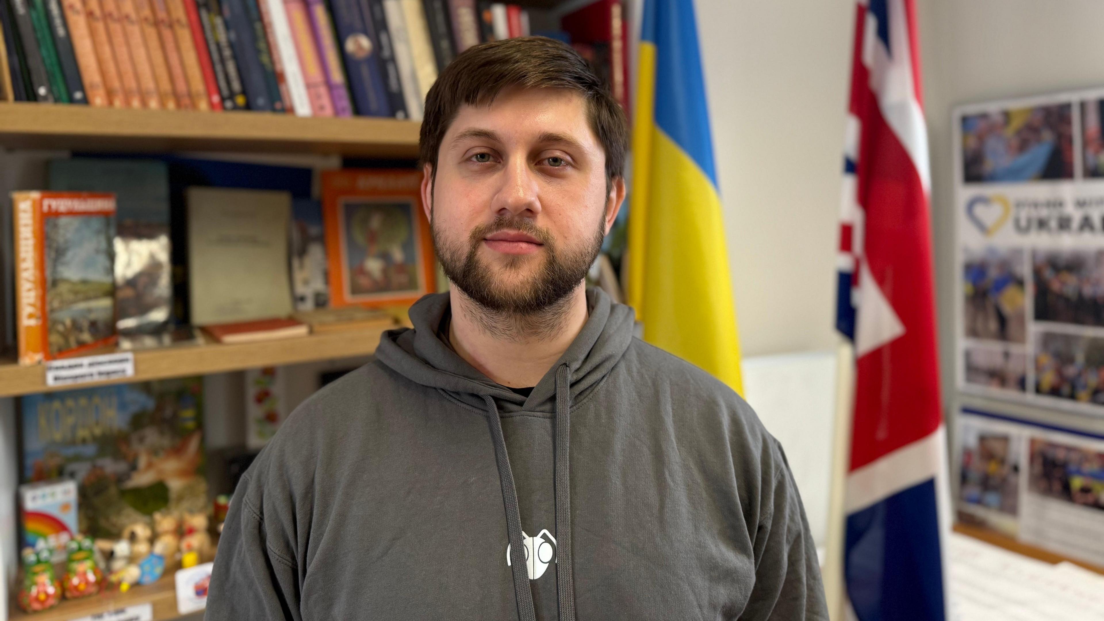 A man with dark hair and stubble wearing a grey hoodie. He's looking straight at the camera and there's a bookshelf, a Ukrainian flag and a Union Jack behind him.