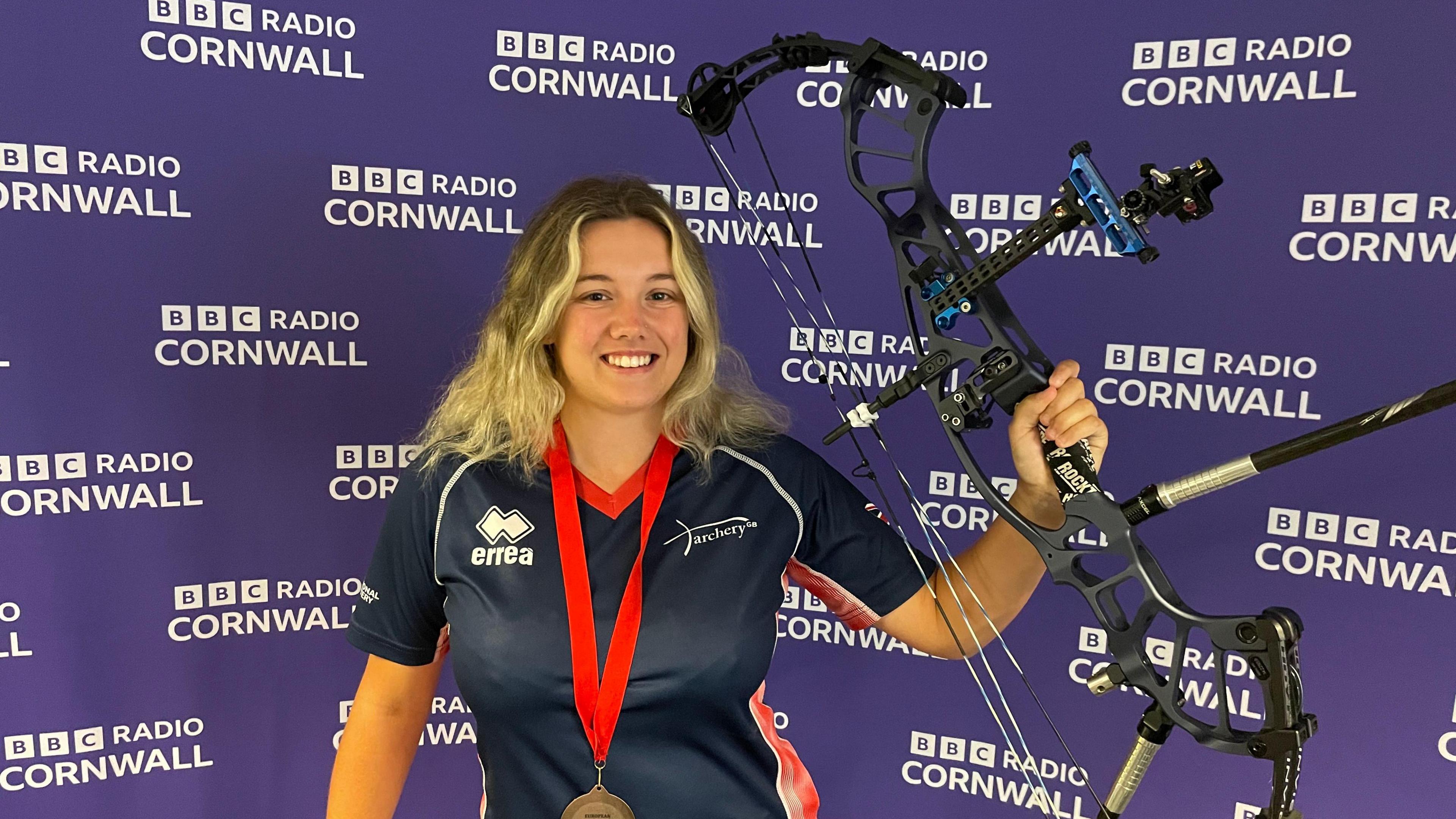 Chloe is smiling at the camera holding her compound bow up high. She is wearing her GB archery uniform and a bronze medal with a red ribbon. She has shoulder-length blonde hair. The purple backdrop is covered with repeated white BBC Radio Cornwall logos.