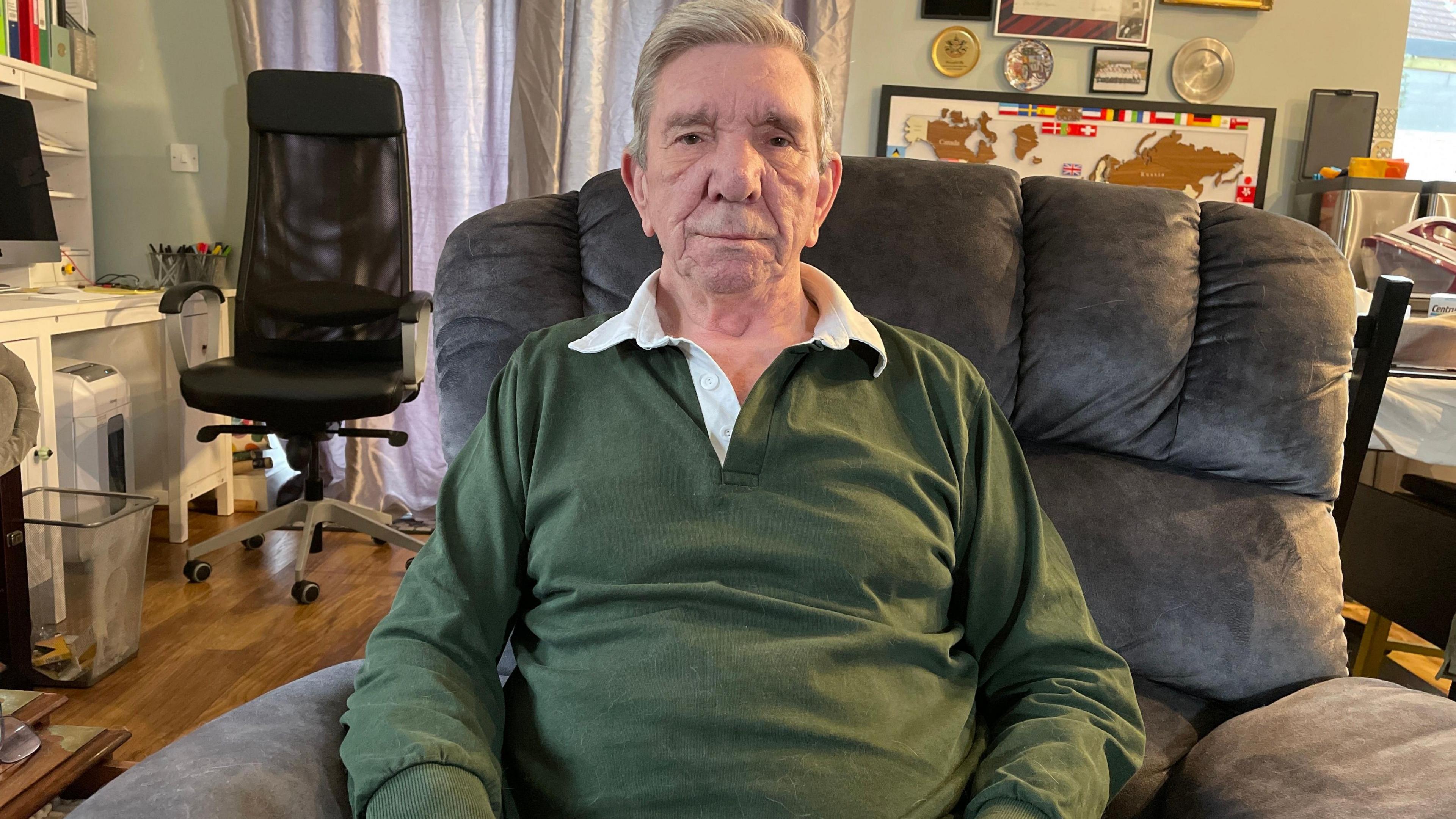 John Heath is wearing a green top and sitting on his sofa