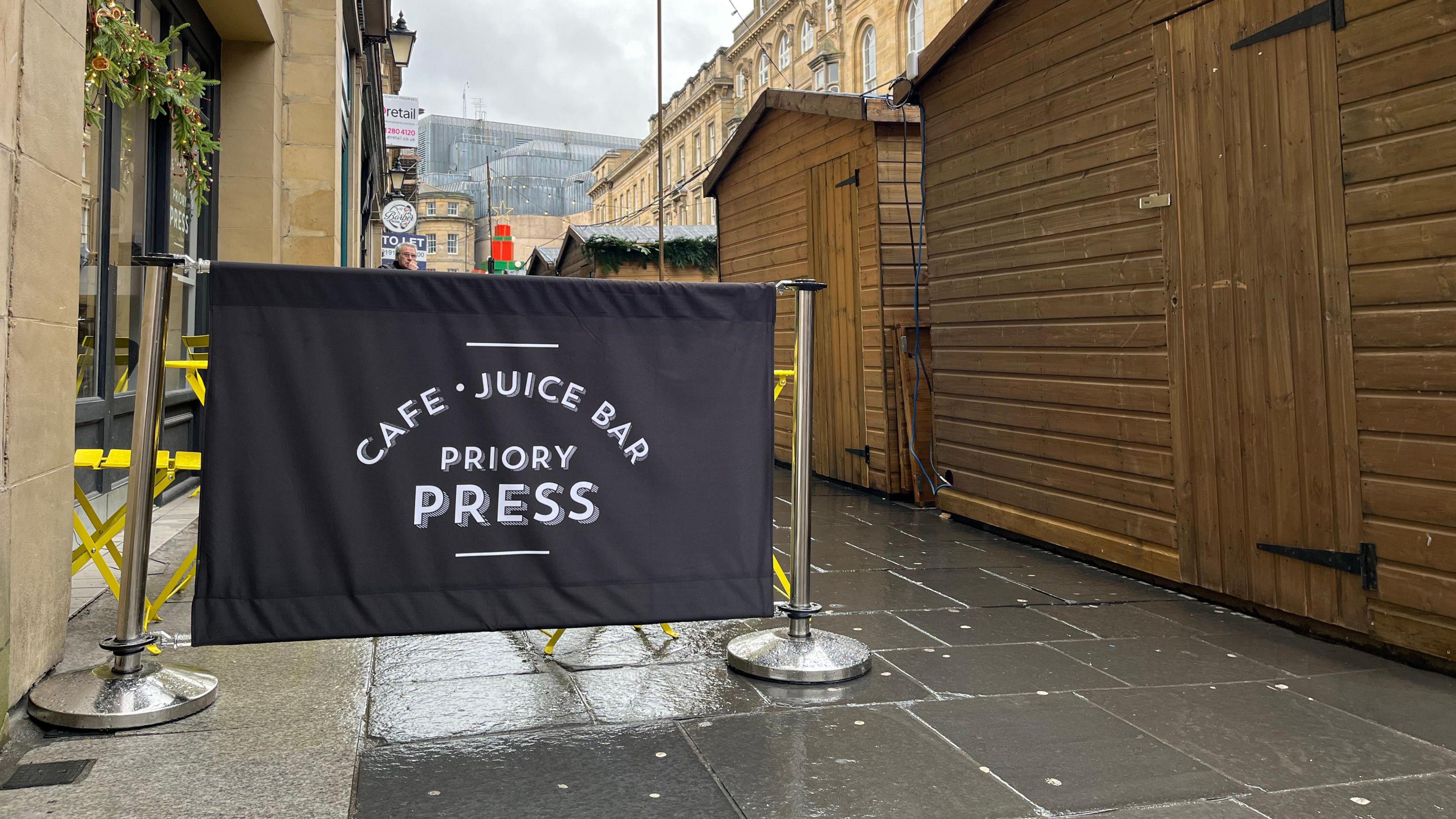 The image shows the external seating area of Priory Press, which is located on the pavement. A short distance away stands a row of several large wooden stalls. Their positioning means the coffee shop now faces the rear wooden walls of the structures.