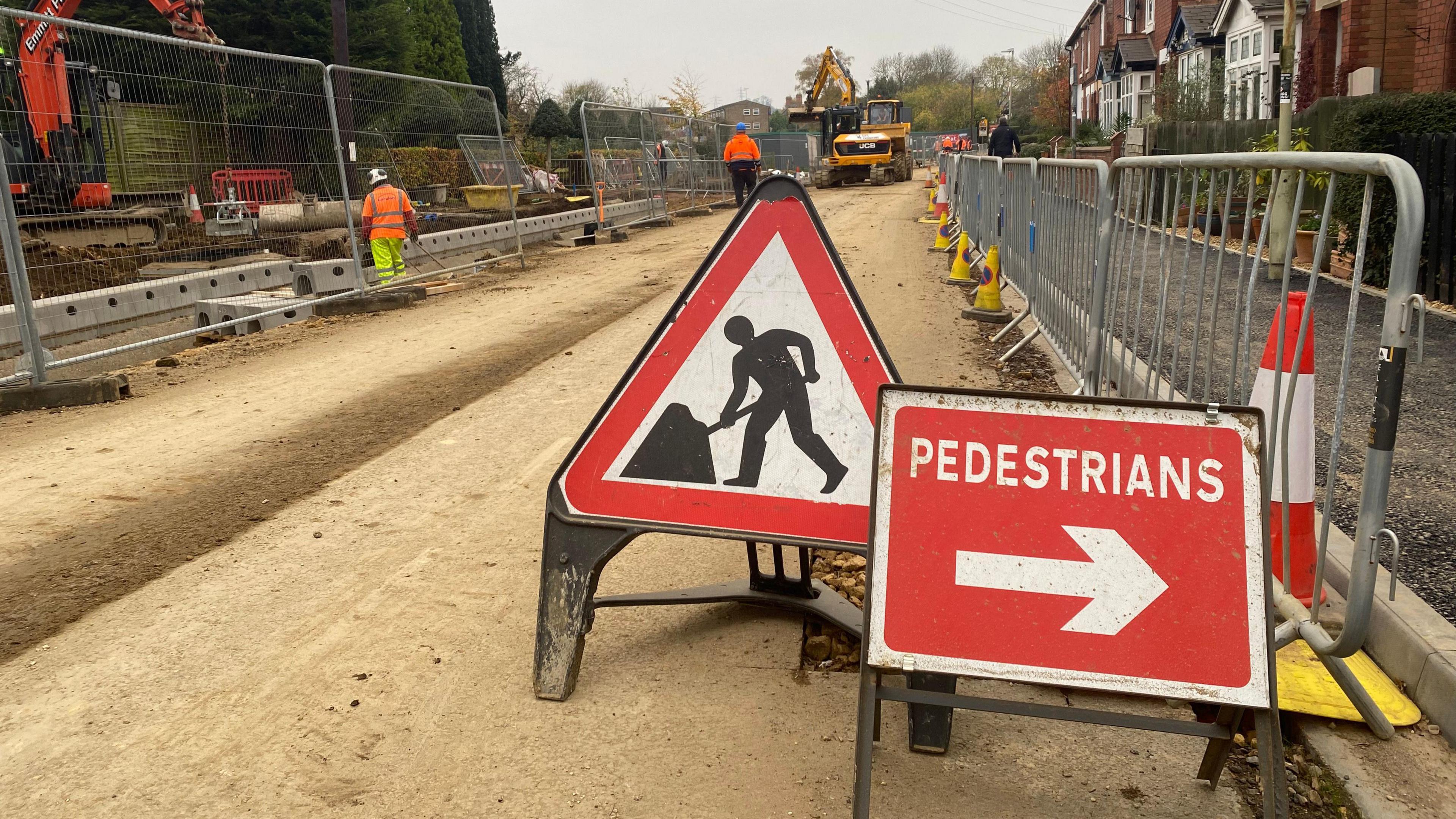 Red road work signs, traffic cones and metal barriers in Brooke Road in Oakham, with a digger in the background.